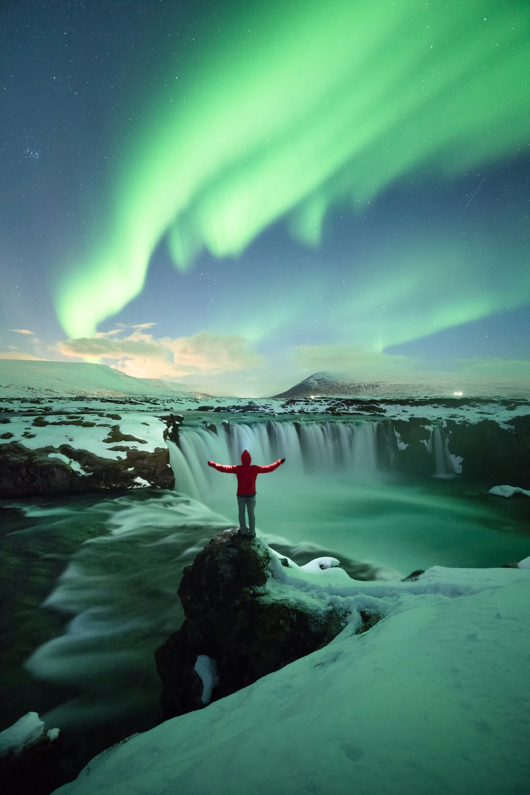 A-person-stands-with-arms-raised-on-a-rock-at-the-top-of-the-horseshoe-shaped-waterfall-Goðafoss-with-green-northern-lights-lighting-up-the-heavens © www.facebook.com / pete.lomchid / Getty Images