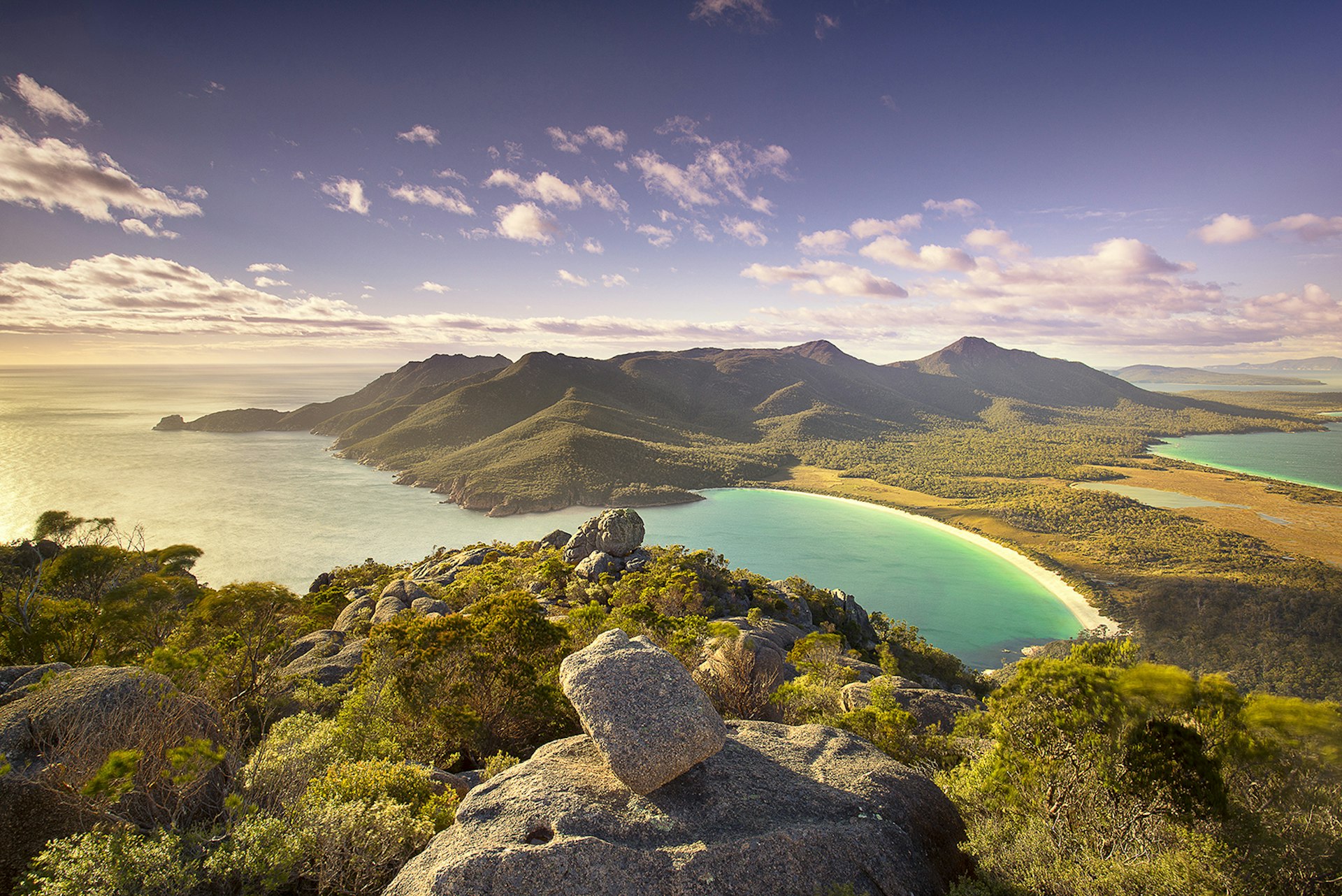 Top of Mt Amos over looking Wineglass Bay, Tasmania at dusk. There are rocks and shrubs in the foreground before giving away to a beautifully smooth beach and blue sea. The sun is low in the sky, throwing out very ambient light. 