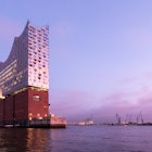 The exterior of Elbphilharmonie (Elbe Philharmonic Hall) in the HafenCity of Hamburg. The photo is taken from the water at dusk. The reflective glass exterior of the concert hall sits on top of a red-brick structure.