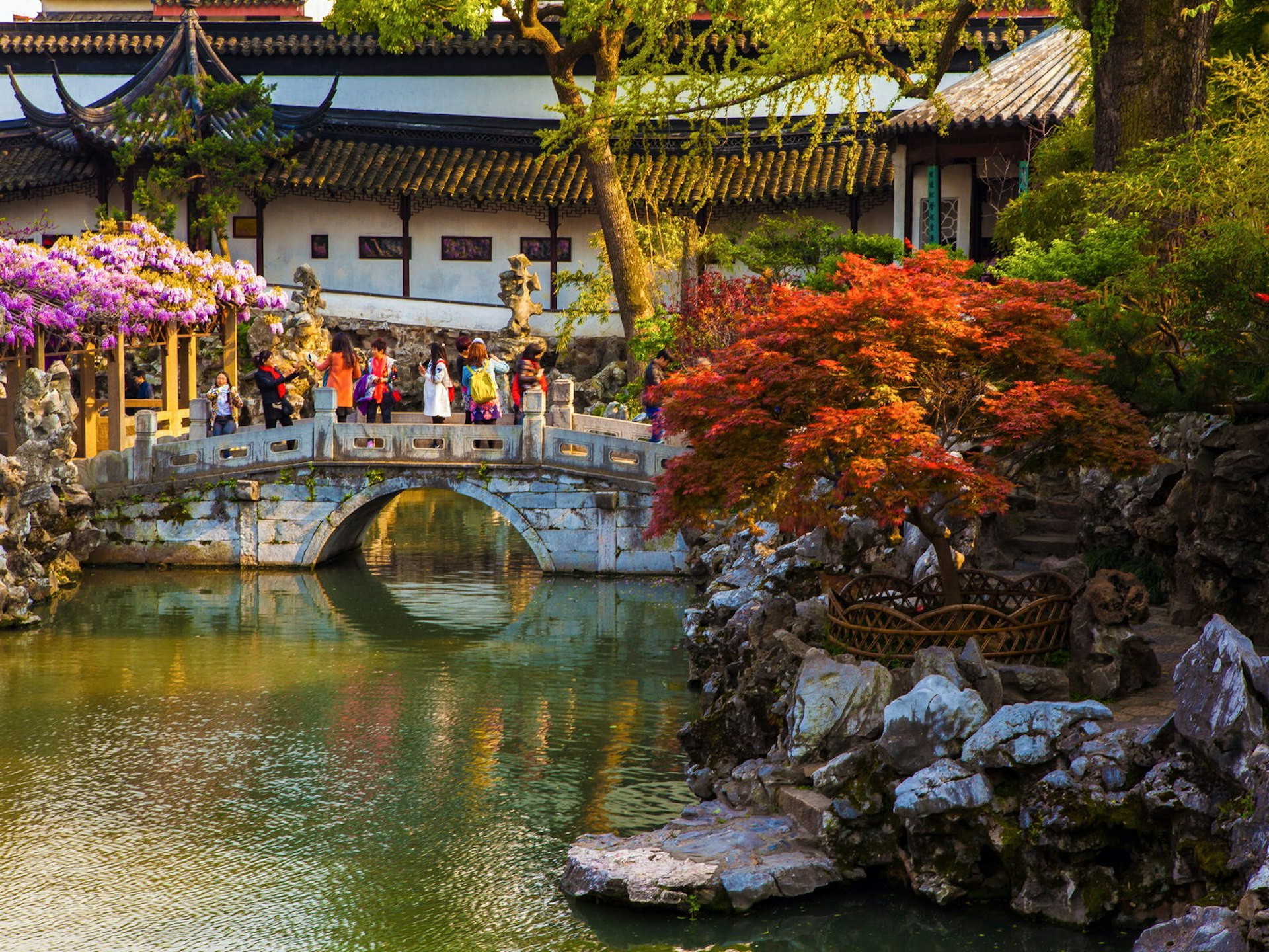 A pond with small stone bridge and covered walkway in the background, framed by red-leafed trees. Classical Chinese landscape architecture – marked by pavilions, covered walkways, rock features and ponds – was developed in Suzhou © Meiqianbao / Shutterstock