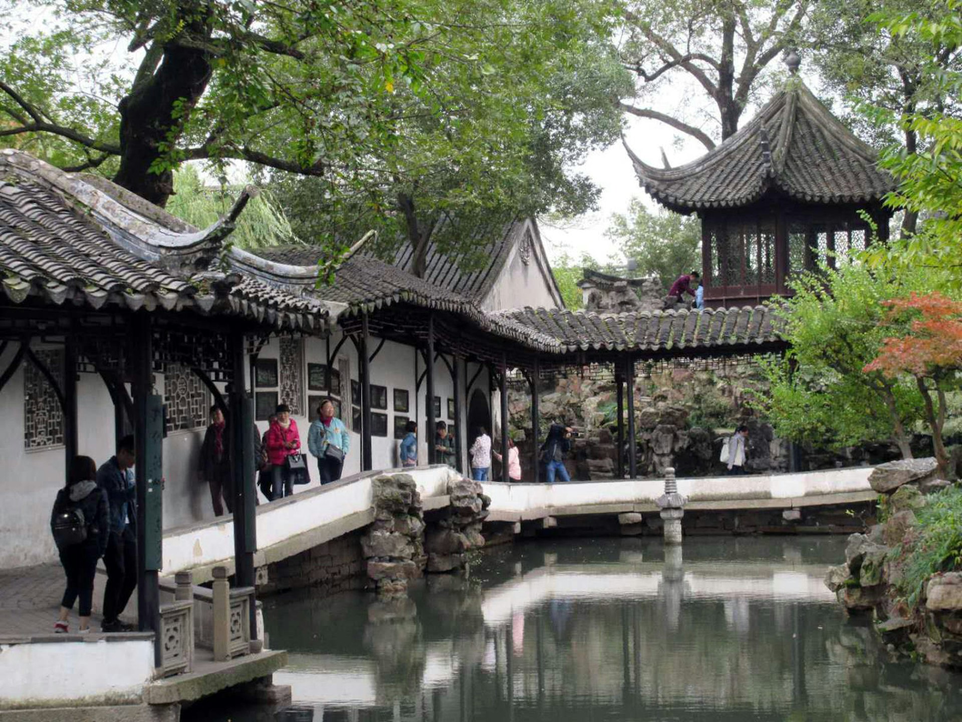 A covered walkway with upturned eaves next to a pond with Chinese pavilion in the background. Suzhou's gardens were first cultivated as private residences © Tess Humphrys / Lonely Planet