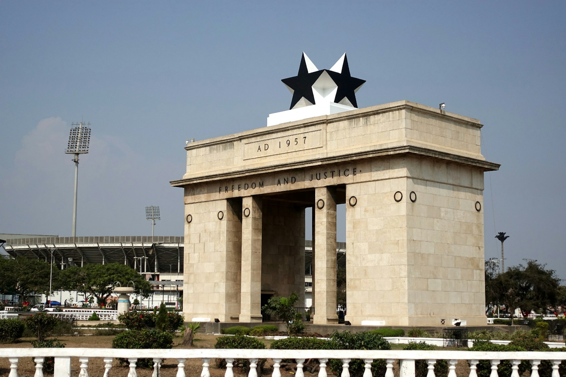 A large, rectangular stone gate, topped by large black stars, stands tall over Independence Square in Accra. The football stadium looms in the distance © Elio Stamm / Lonely Planet
