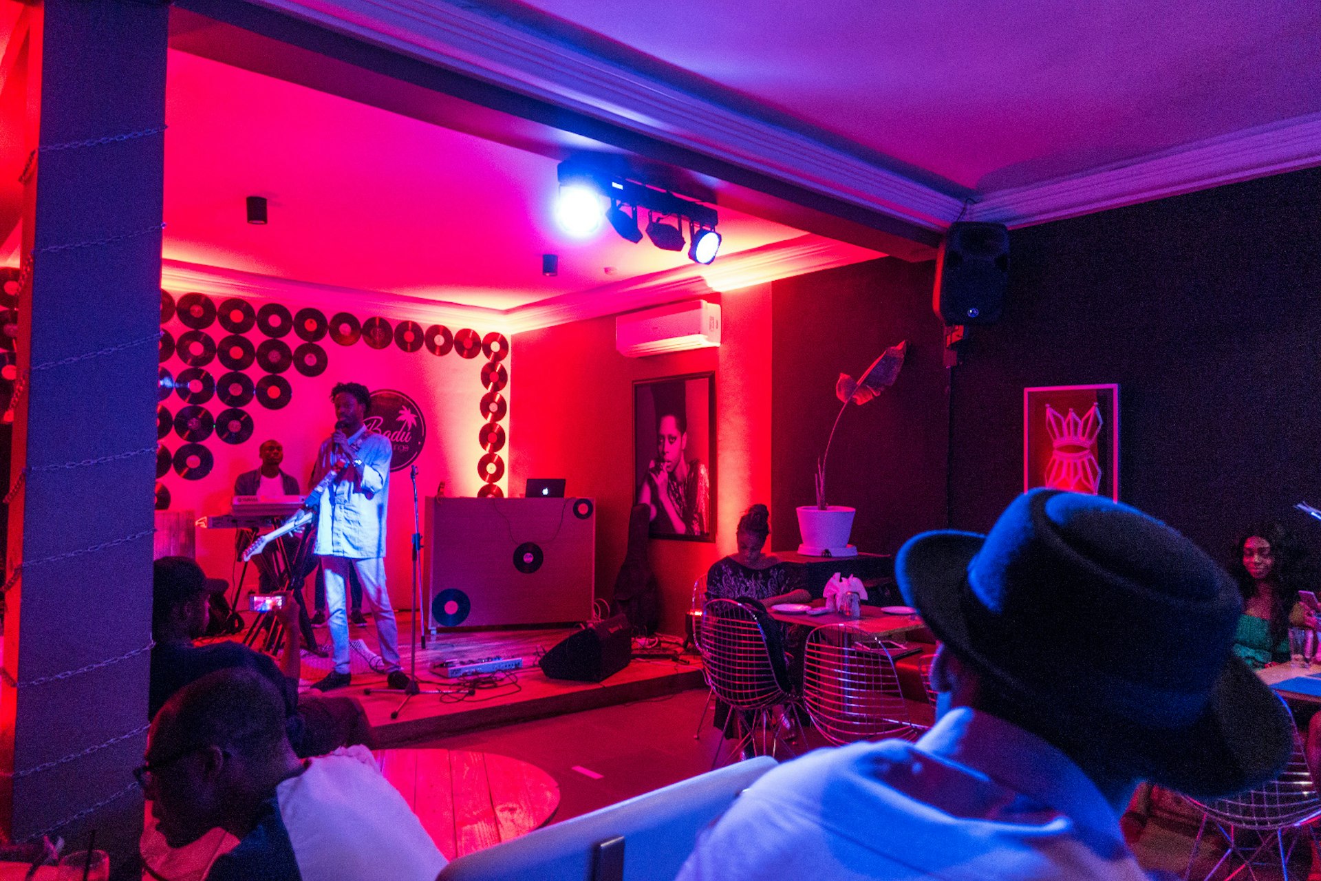 A small stage and room glow red, with a singer on stage holding a guitar and microphone. The back wall of the stage is decorated with a pattern of old vinyl records © Elio Stamm / Lonely Planet