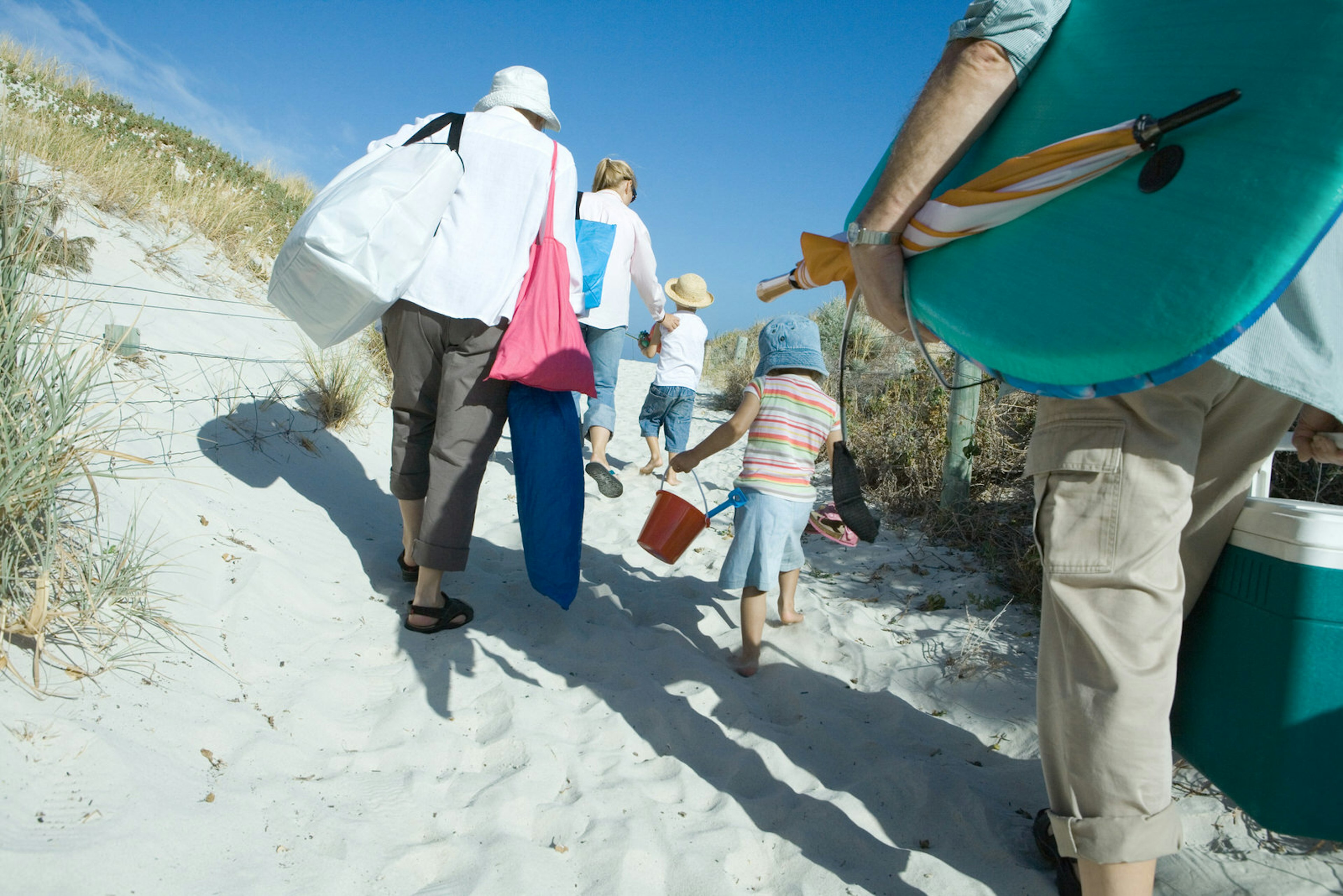 Rear view of a family group of three adults and two young children, make their way through sand dunes carrying a lot of beach items