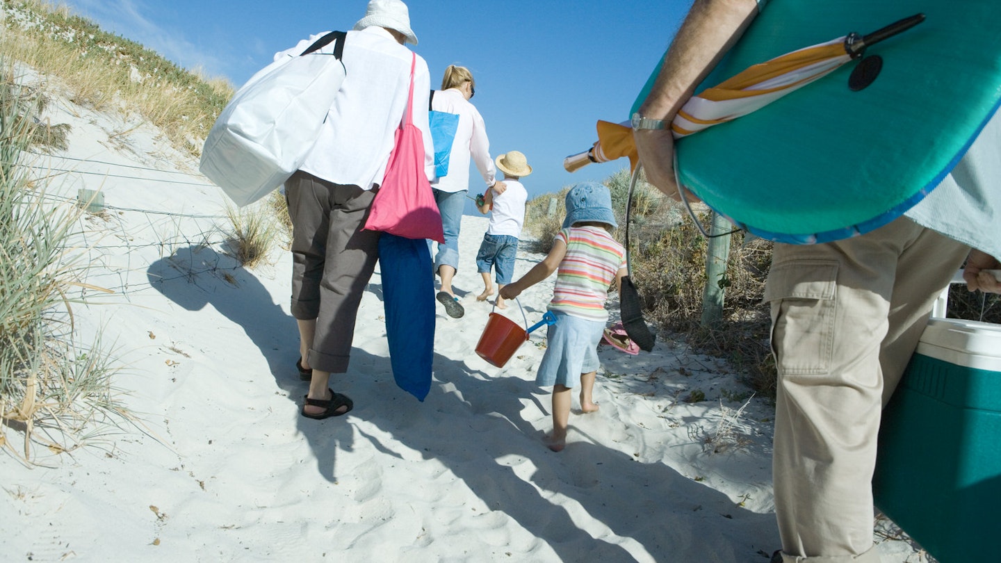 Rear view of a family group of three adults and two young children, make their way through sand dunes carrying a lot of beach items