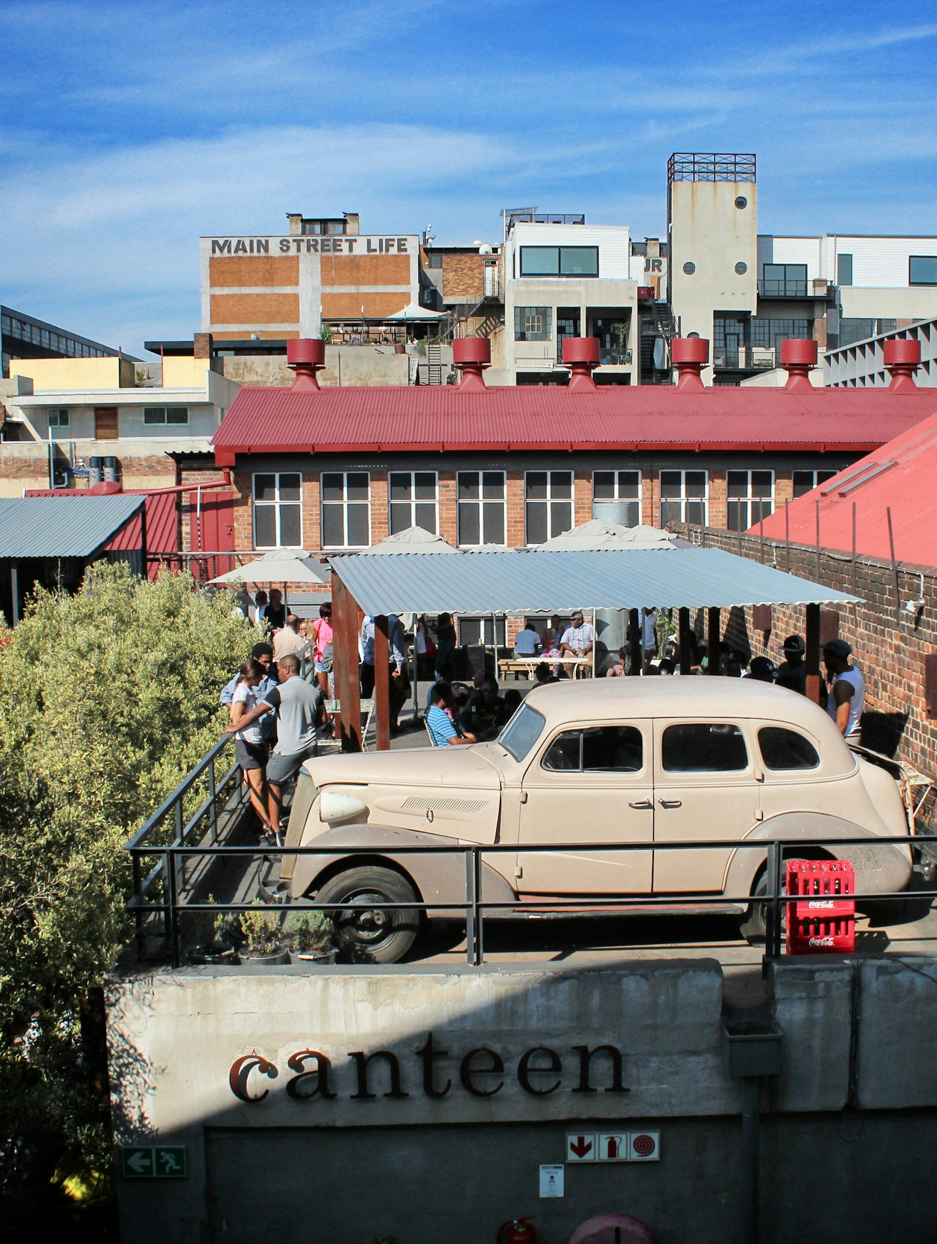 A crowd gathers under an awning on the flat roof of Market on Main, sharing the space is a vintage 1930s car. Trees climb up to this level on the left and renovated old industrial buildings back the scene ©Antonella Ragazzoni / 500px