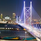Stretching over eight rows of parked trains, the brilliantly lit white towers and cables of the Nelson Mandela Bridge stretch towards the skyline © Henrique NDR Martins / Getty Images
