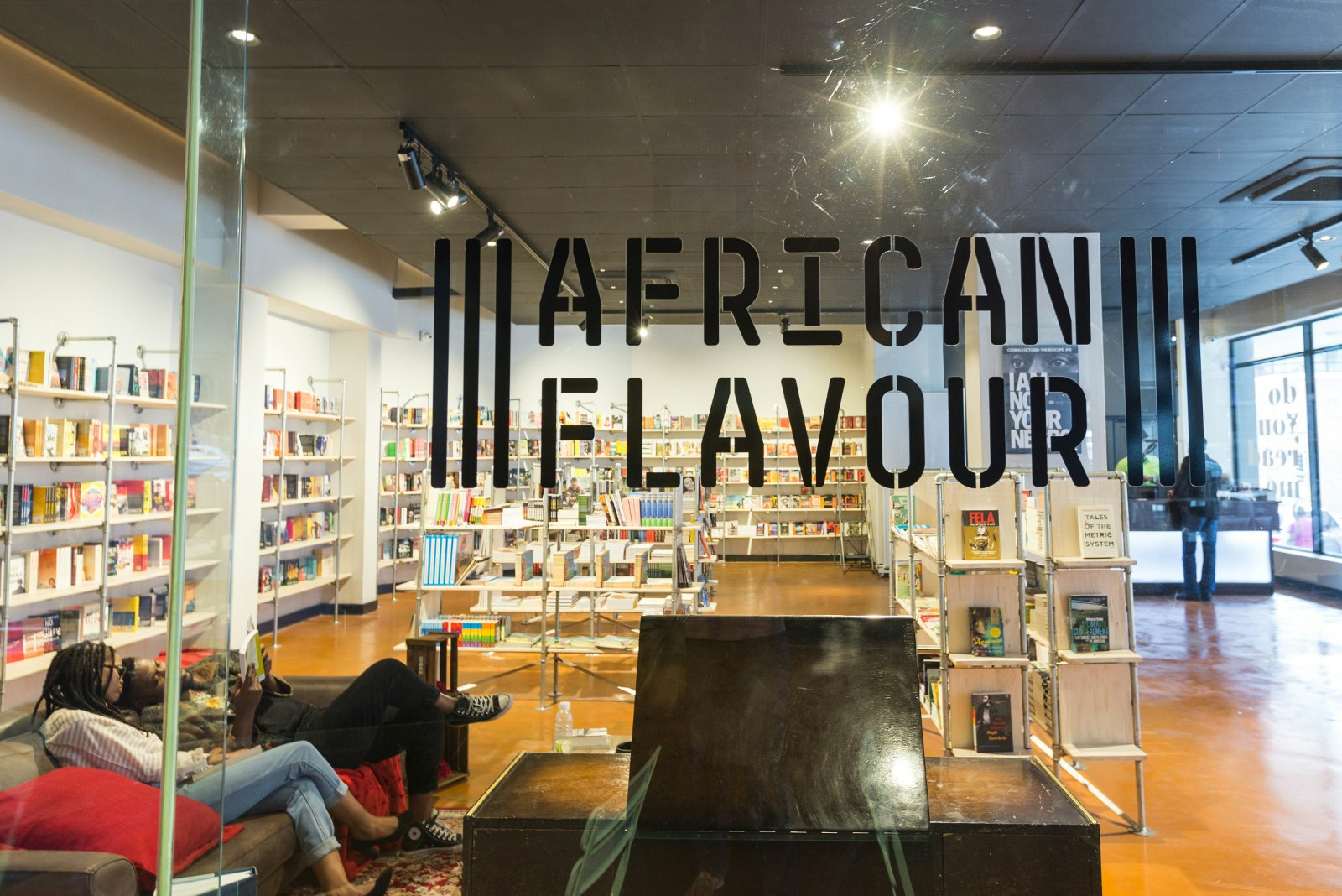 Behind a glass wall emblazoned with black letters reading African Flavour is a wood-floored room with white walls and white bookshelves filled with colourful books. Patrons sit on sofas reading © Heather Mason / Lonely Planet