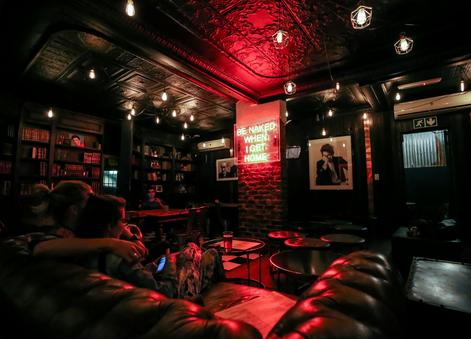 A couple sit in a leather chesterfield sofa in this darkened bar, with a red glow radiating across the ceiling, tables and sofa from a neon sign reading 'Be naked when I get home' - next to the sing is a framed black-and-white poster of Bob Dylan © Heather Mason / Lonely Planet
