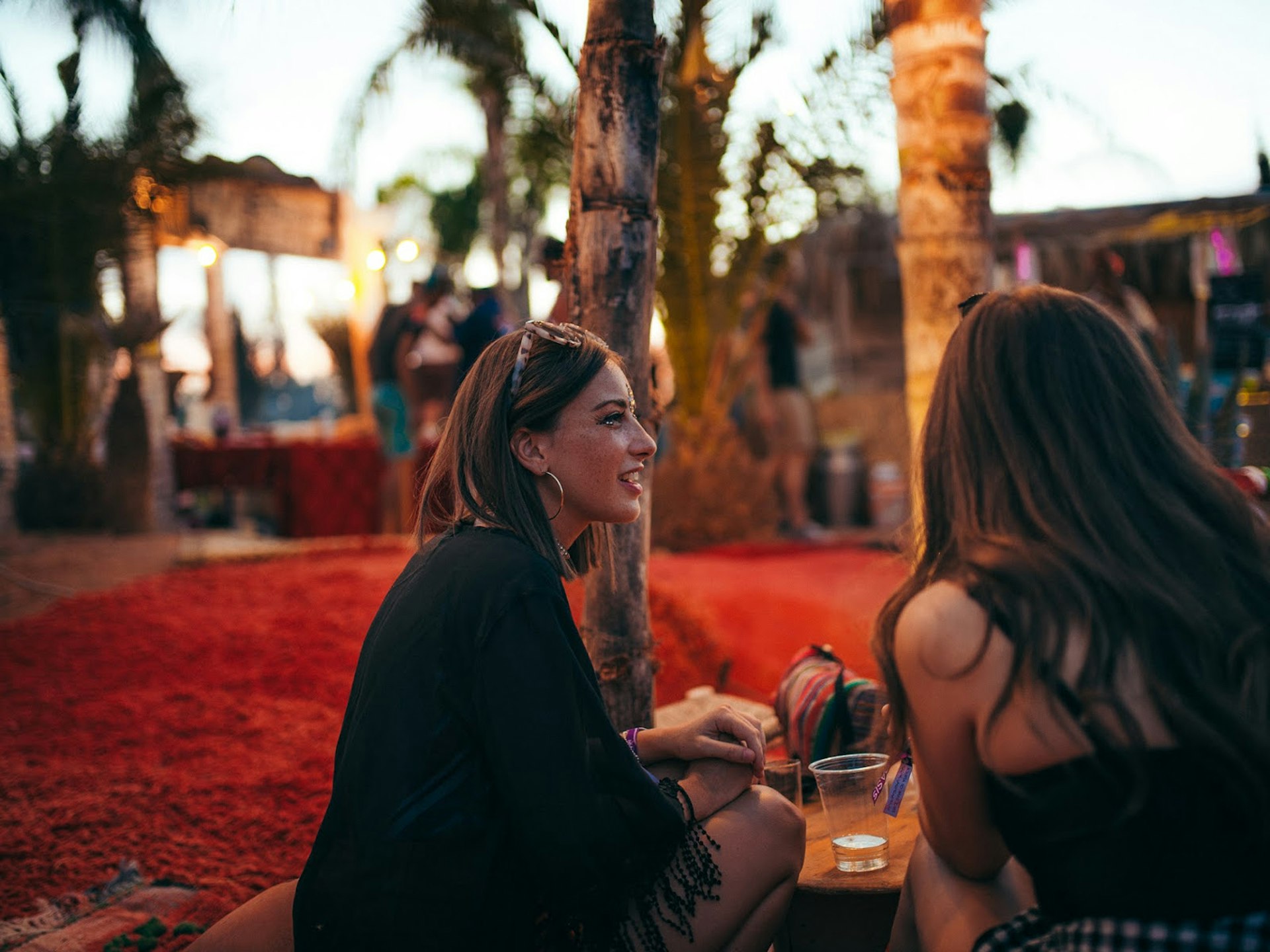 Alternative festivals - two women relaxing sharing drinks at the Oasis Festival, Morocco at dusk. The floor appears to be made of red sand and there are palm trees and twinkling lights in the background.
