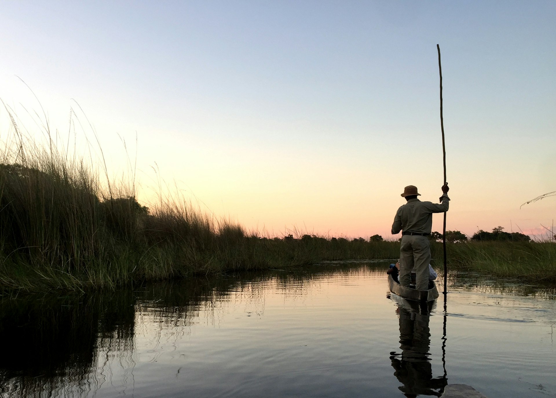 A poler stands at the back of a mokoro (dugout canoe) with his pole raised vertically to the sky. His outline is crisp against a pinkish blue sky at sunset. To his left are a bed of reeds at the edge of the channel. Everything is reflected in the still water of the delta © Matt Phillips / Lonely Planet