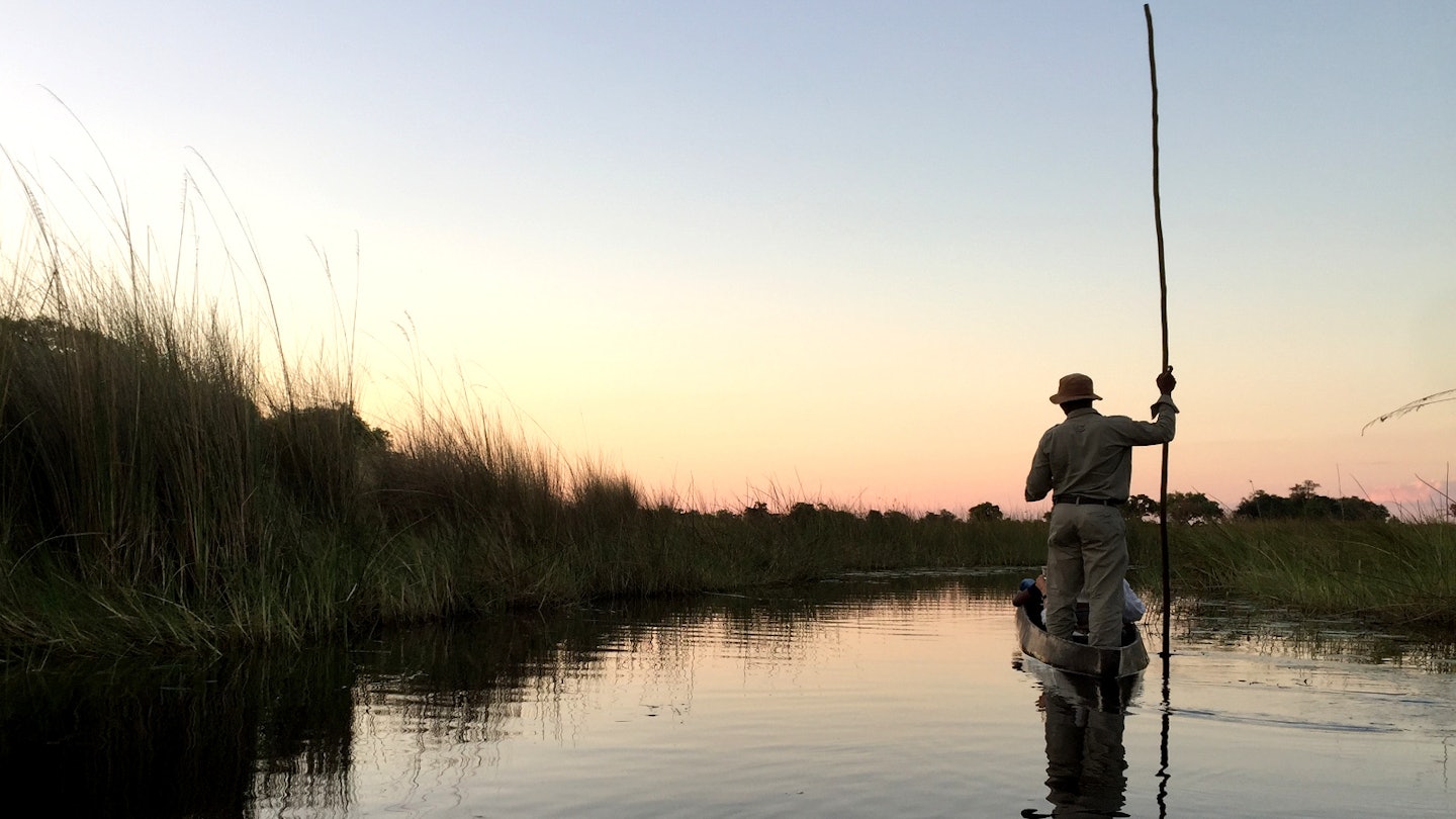 A poler stands at the back of a mokoro (dugout canoe) with his pole raised vertically to the sky. His outline is crisp against a pinkish blue sky at sunset. To his left are a bed of reeds at the edge of the channel. Everything is reflected in the still water of the delta © Matt Phillips / Lonely Planet
