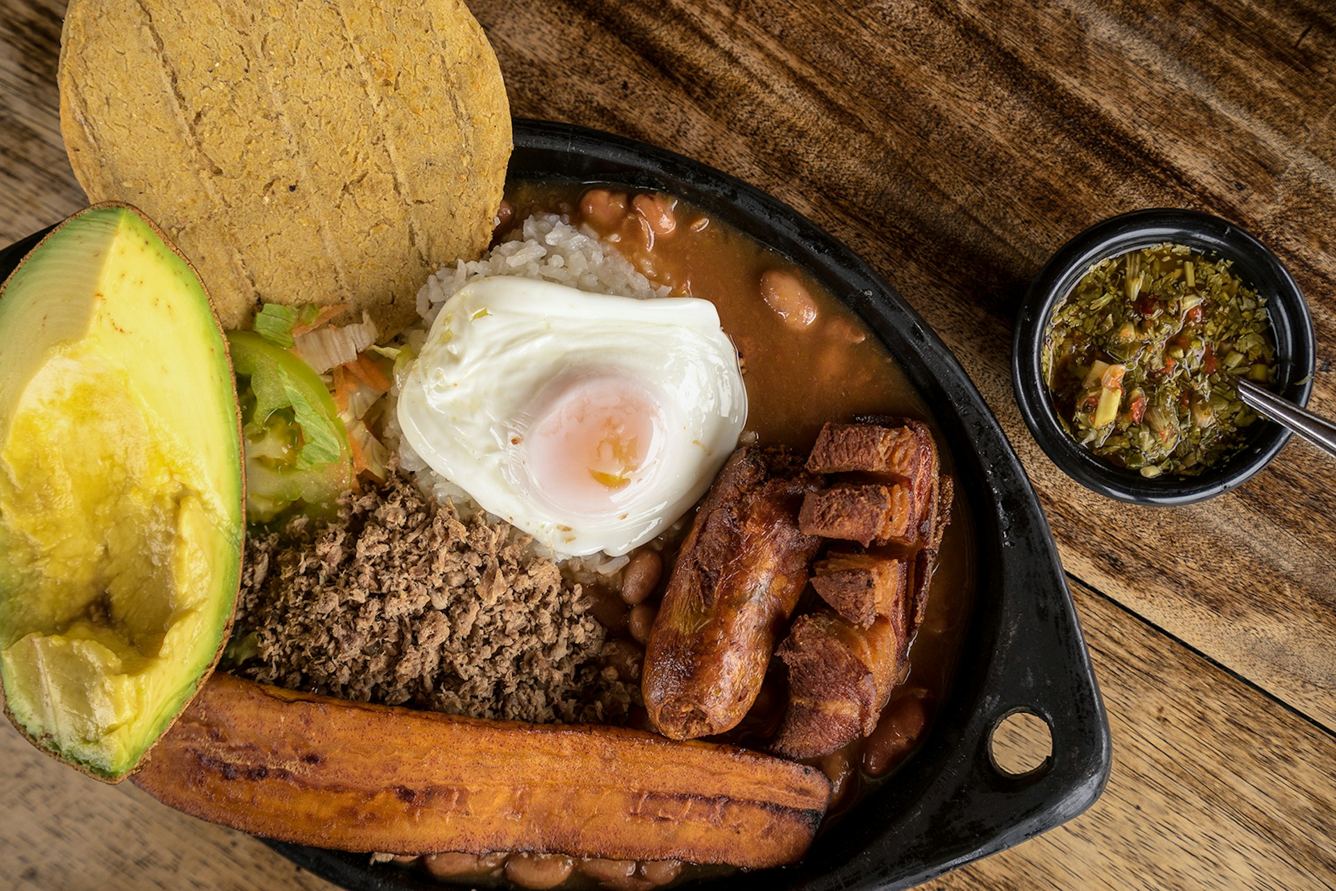 A plate of food with an egg, avocado, plantains, ground beef, arepas, beans and sausage.