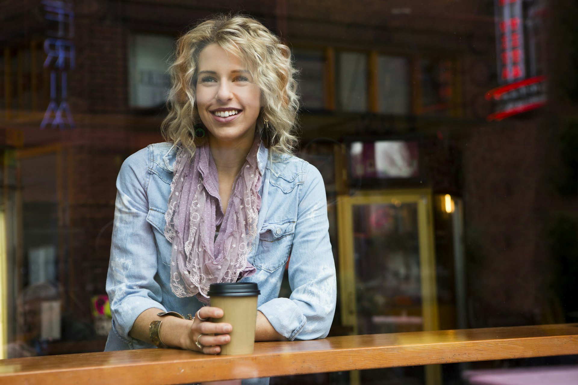 An attractive young woman drinks coffee from a paper cup in Portland. A reflection of other nearby buildings is shown in the window © Jordan Siemens / Getty Images