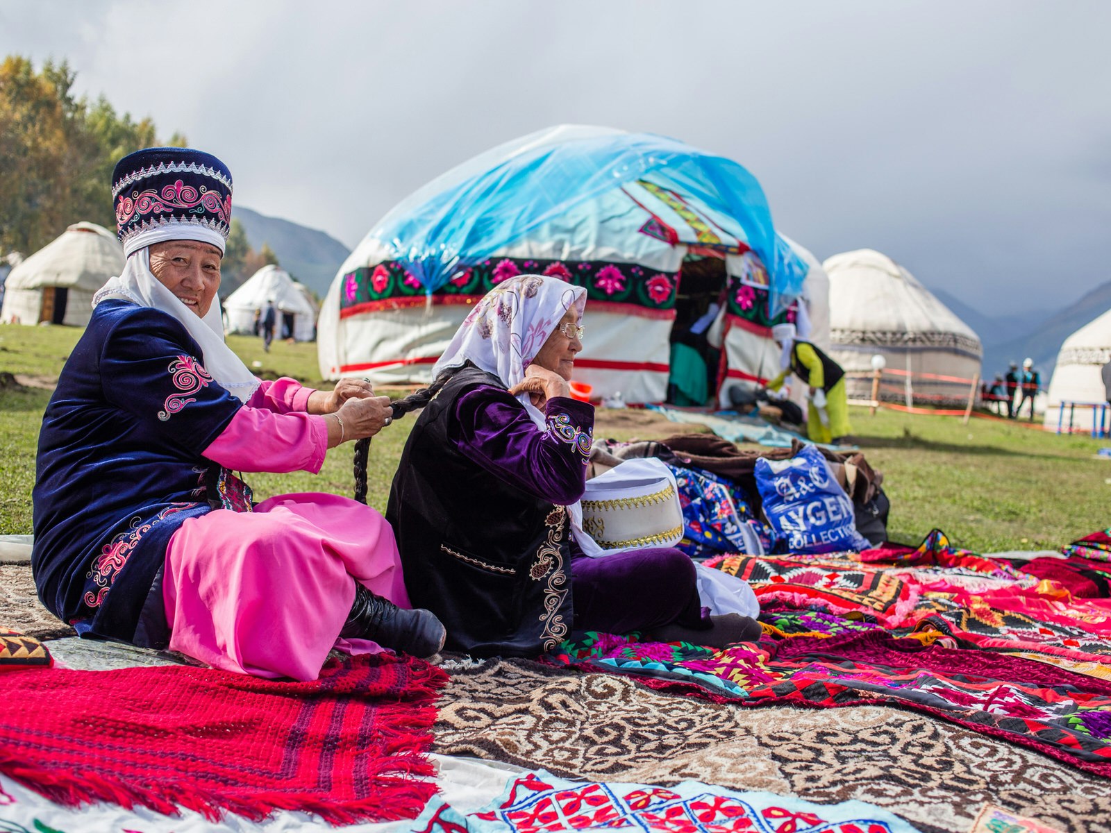 Two elderly Kyrgyz ladies in traditional dress sit on colourful blankets on the grass, with a few yurts and mountains in the background. Many of the World Nomad Games events take place here, in the Kyrchyn valley, where the yurt camp offers accommodation and traditional activities © Stephen Lioy / Lonely Planet