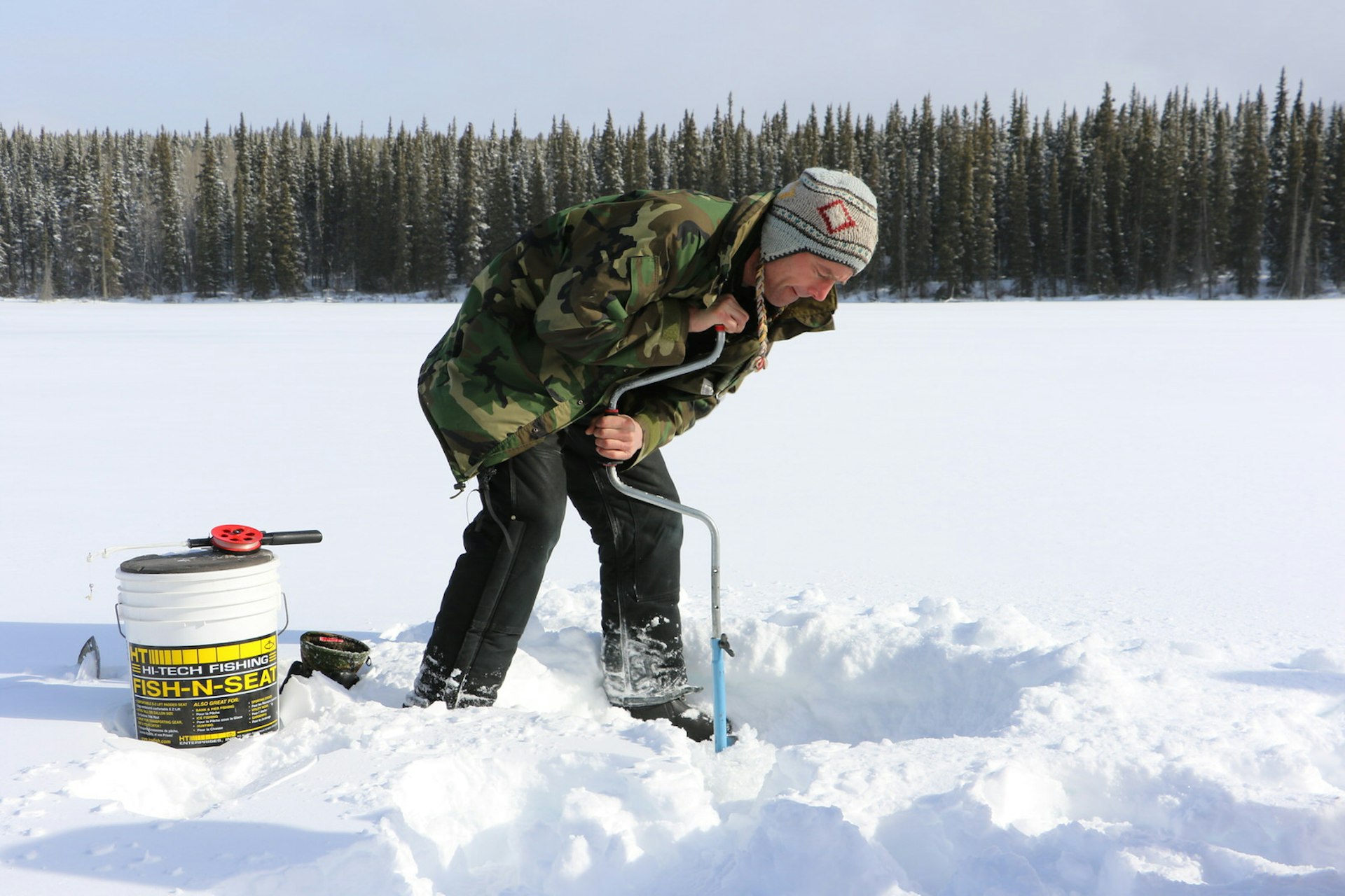 A man uses a hand-powered drill to make a hole in a snow- and ice-covered lake. A tackle box is near at hand © Mike MacEacheran / Lonely Planet