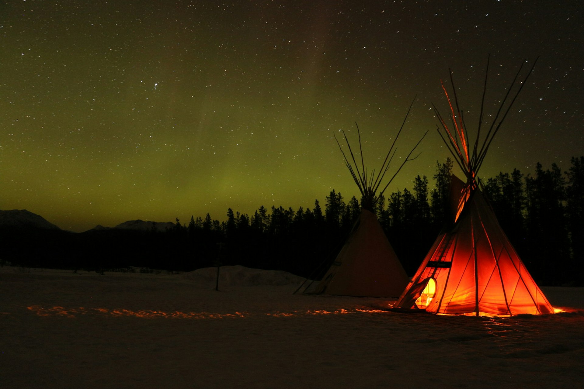 The northern lights glow in the distance on a black landscape while a tipi glows in the foreground © Mike MacEacheran / Lonely Planet