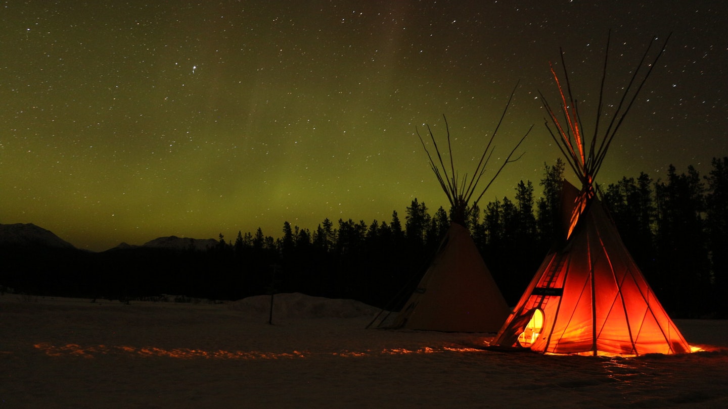 The northern lights glow in the distance on a black landscape while a tipi glows in the foreground © Mike MacEacheran / Lonely Planet