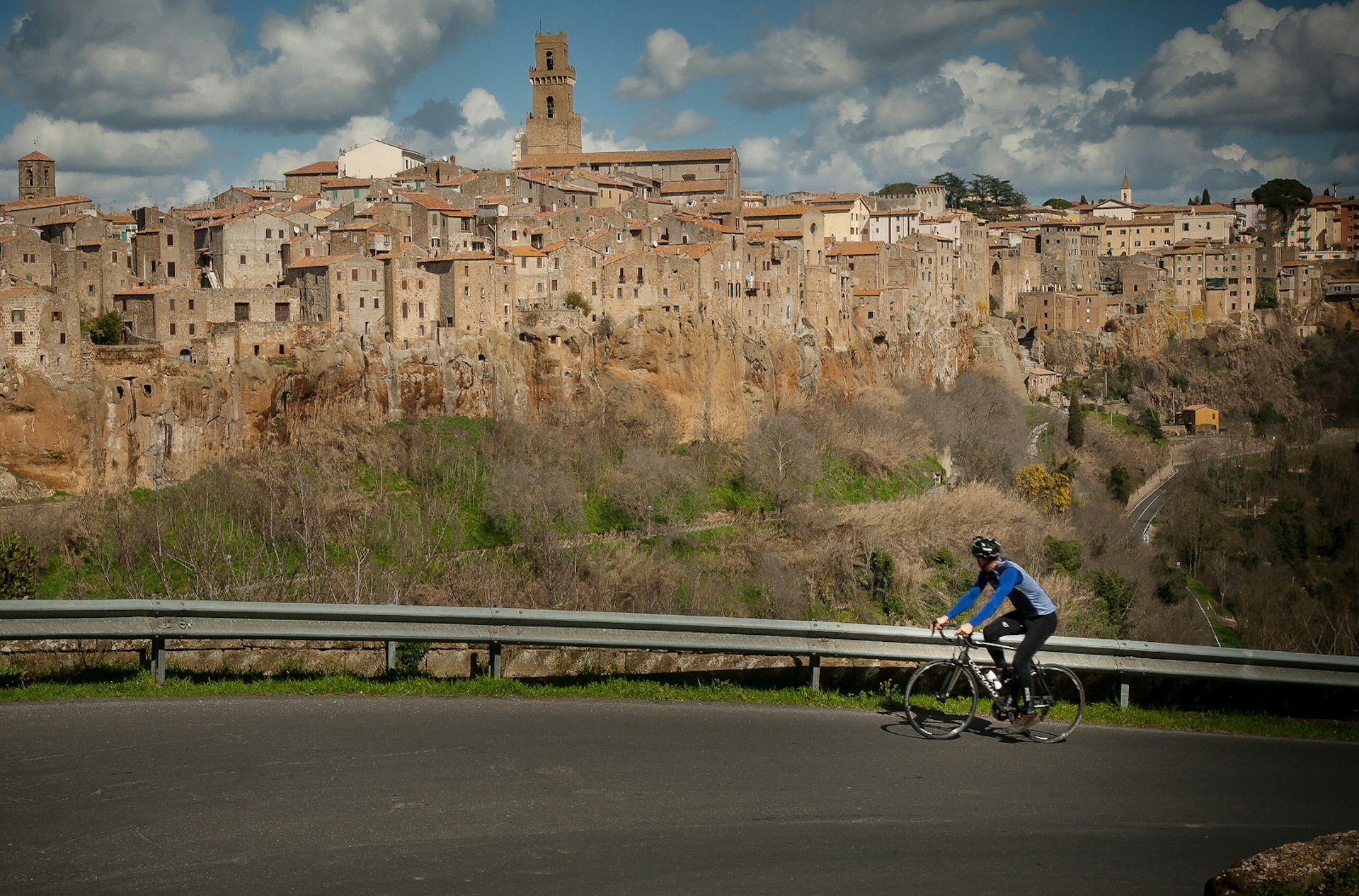 A cyclist climbing up a steep road, with his head turned away from the camera and towards the impressive medieval skyline of Pitigliano © Ciclica & Foto Mariollorca.com