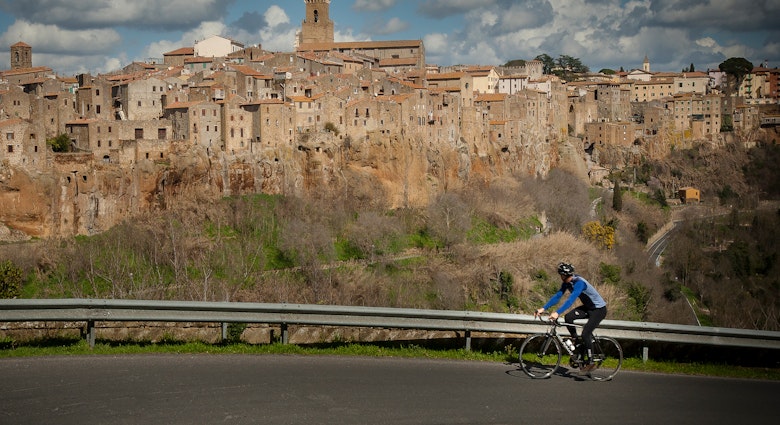 A cyclist climbing up a steep road, with his head turned away from the camera and towards the impressive medieval skyline of Pitigliano © Ciclica & Foto Mariollorca.com