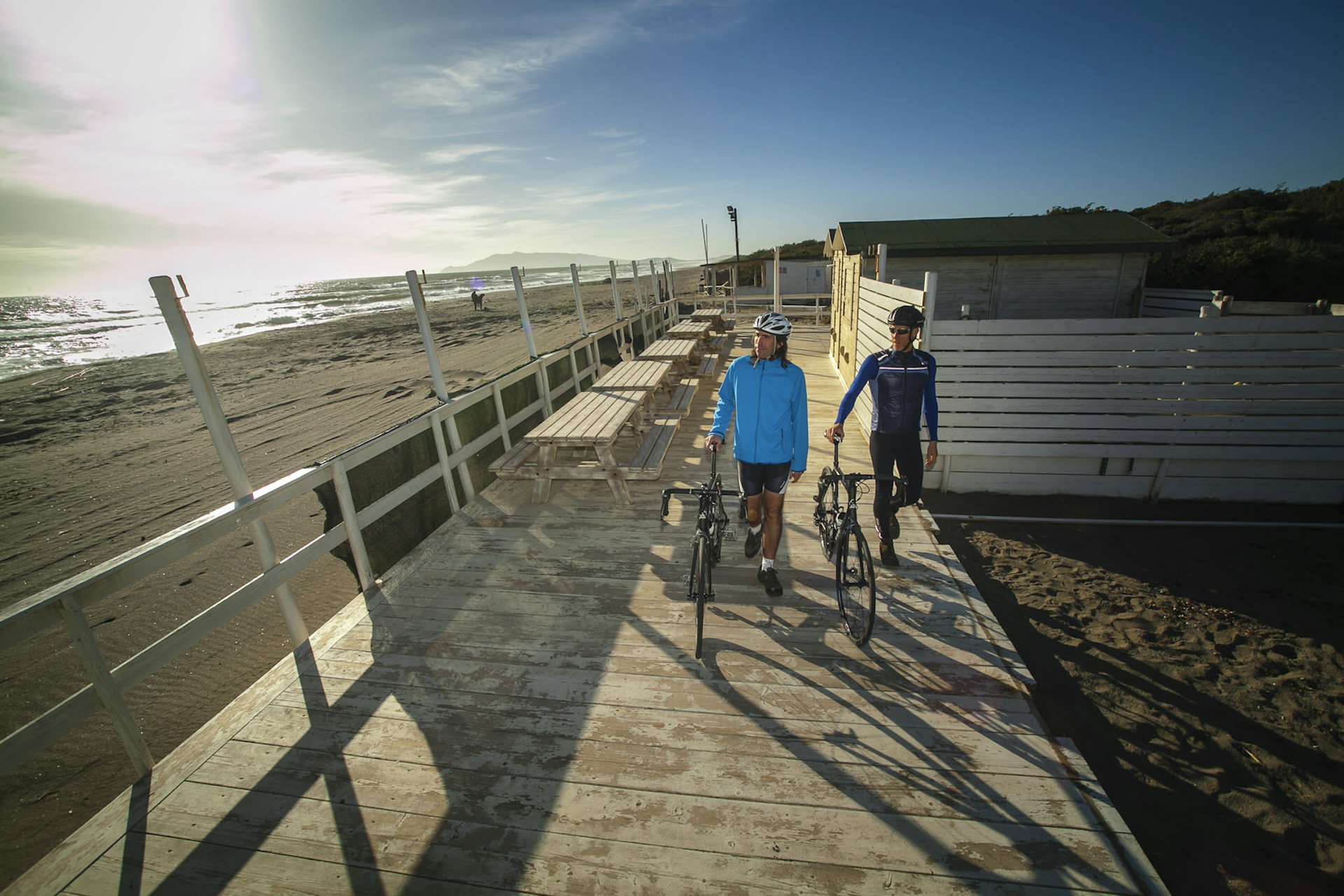Two cyclists walk their bikes along the boardwalk that backs an endless section of beach at Chiarone Scalo © Ciclica & Foto Mariollorca.com