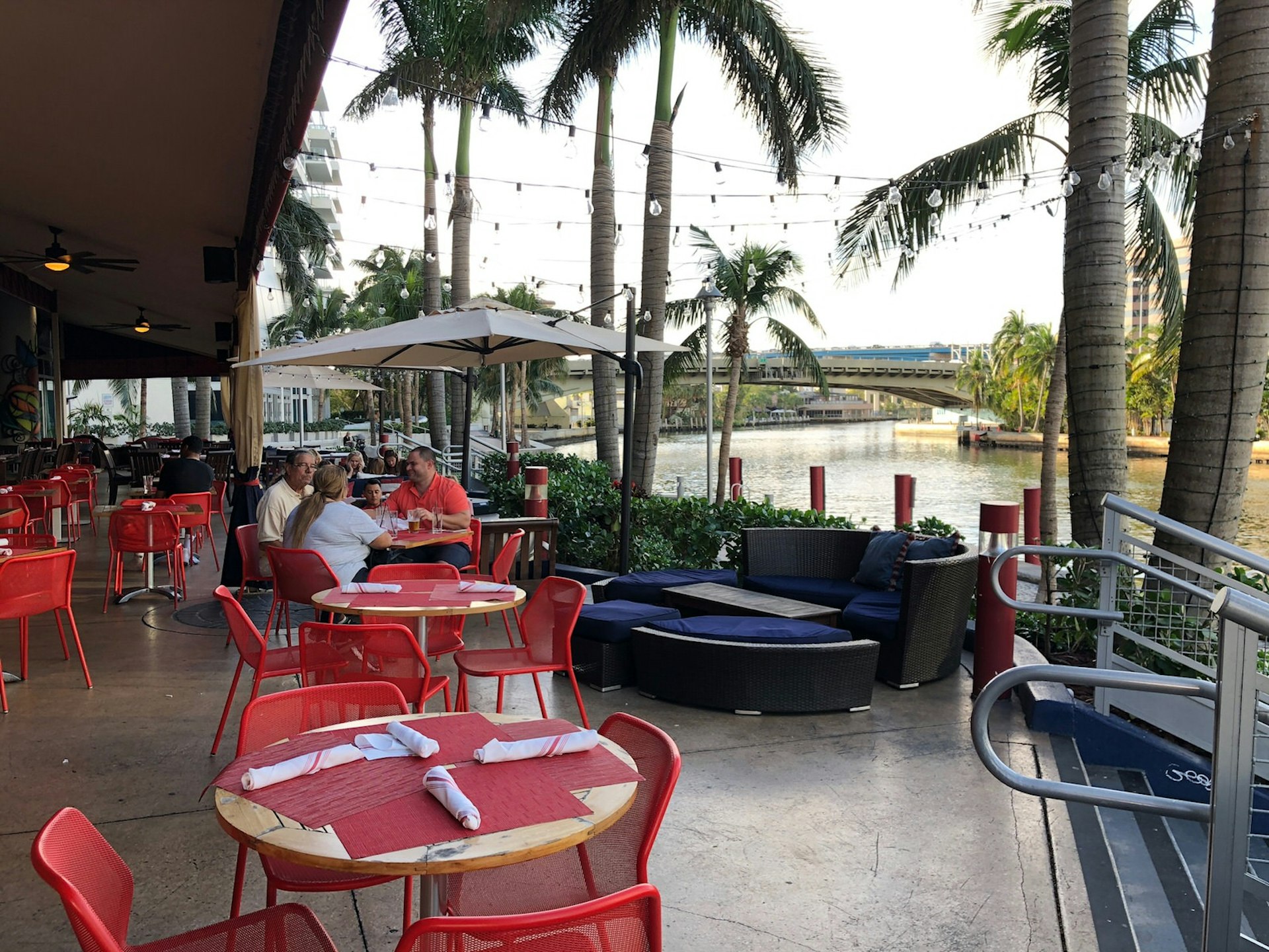 Red tablecloths lend a pop of color to a waterfront bar area with palm trees and sand beyond © Jackie Gutierrez-Jones / Lonely Planet