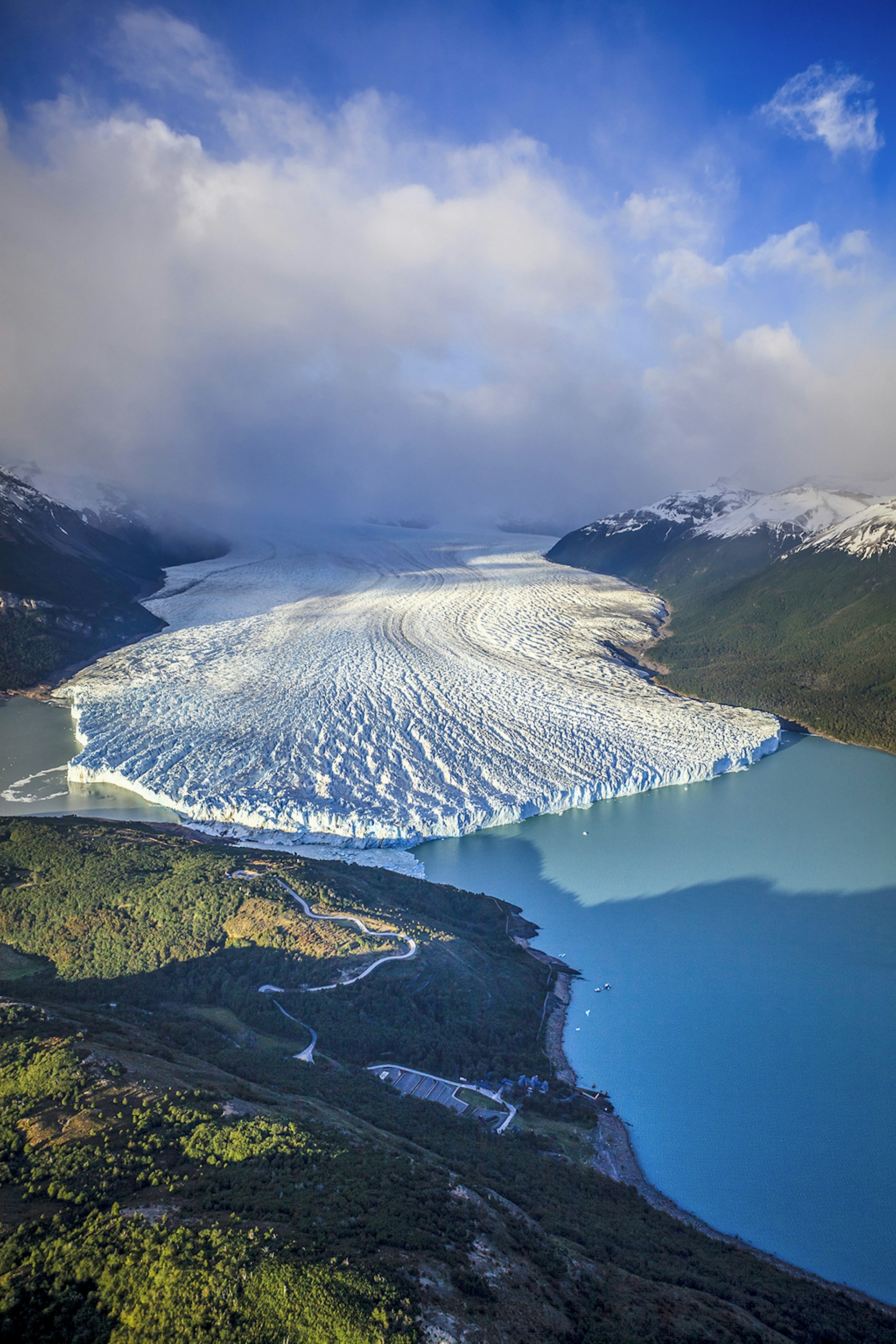 An aerial view of a glacial tongue extending into a sky-blue lake