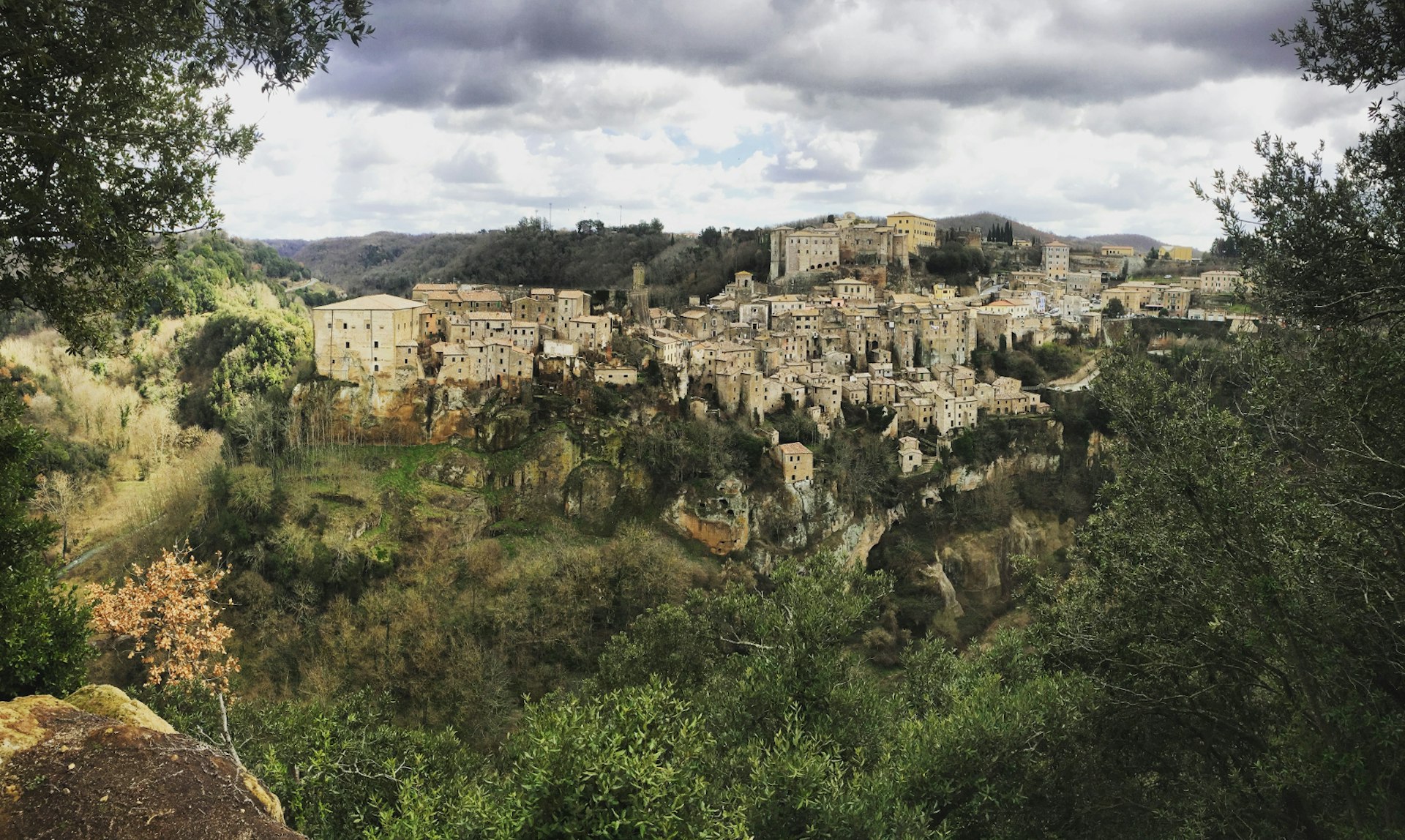 The image is looking over a green, forested gorge to the grey stone buildings of Sorano protruding from the trees and poking into a cloudy sky © Matt Phillips / Lonely Planet