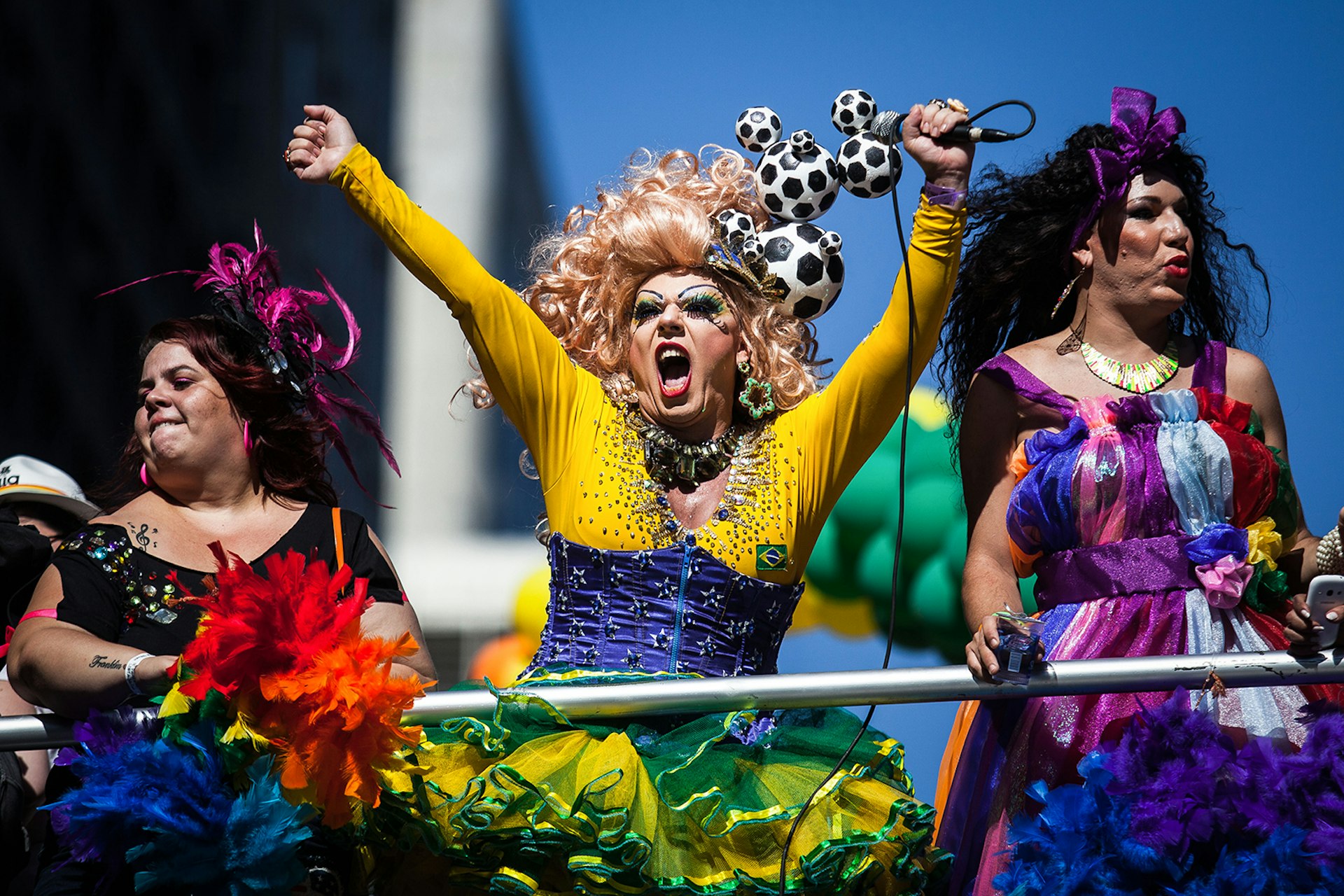 Three brightly clothed individuals celebrate Pride in Sao Paulo; one is wearing a Brazil soccer themed dress © Victor Moriyama/Getty Images