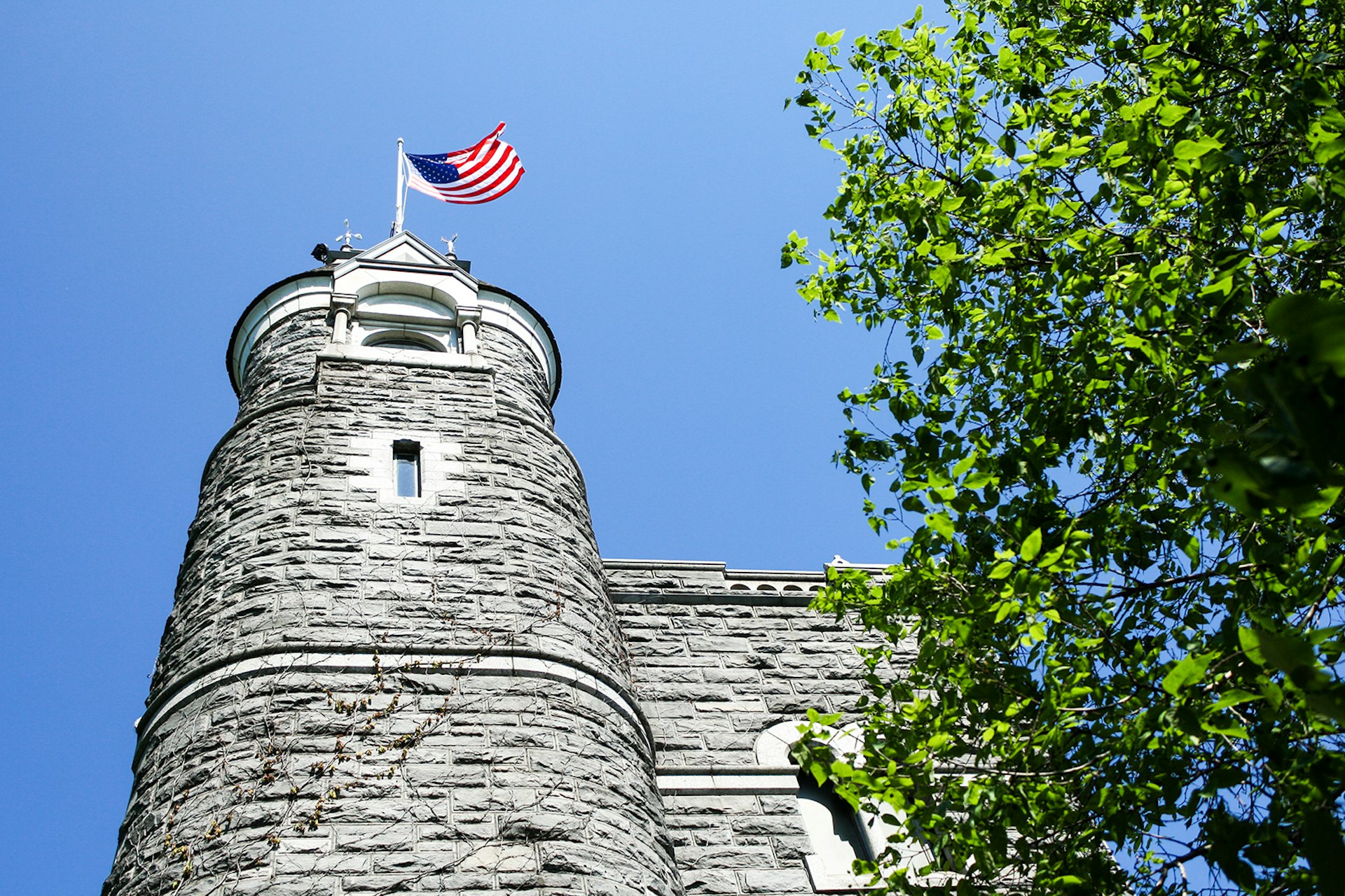 A gray stone tower topped by an American flag extends into the blue sky, framed by a green, leafy tree, in Central Park © Brian Bumby / Getty Images