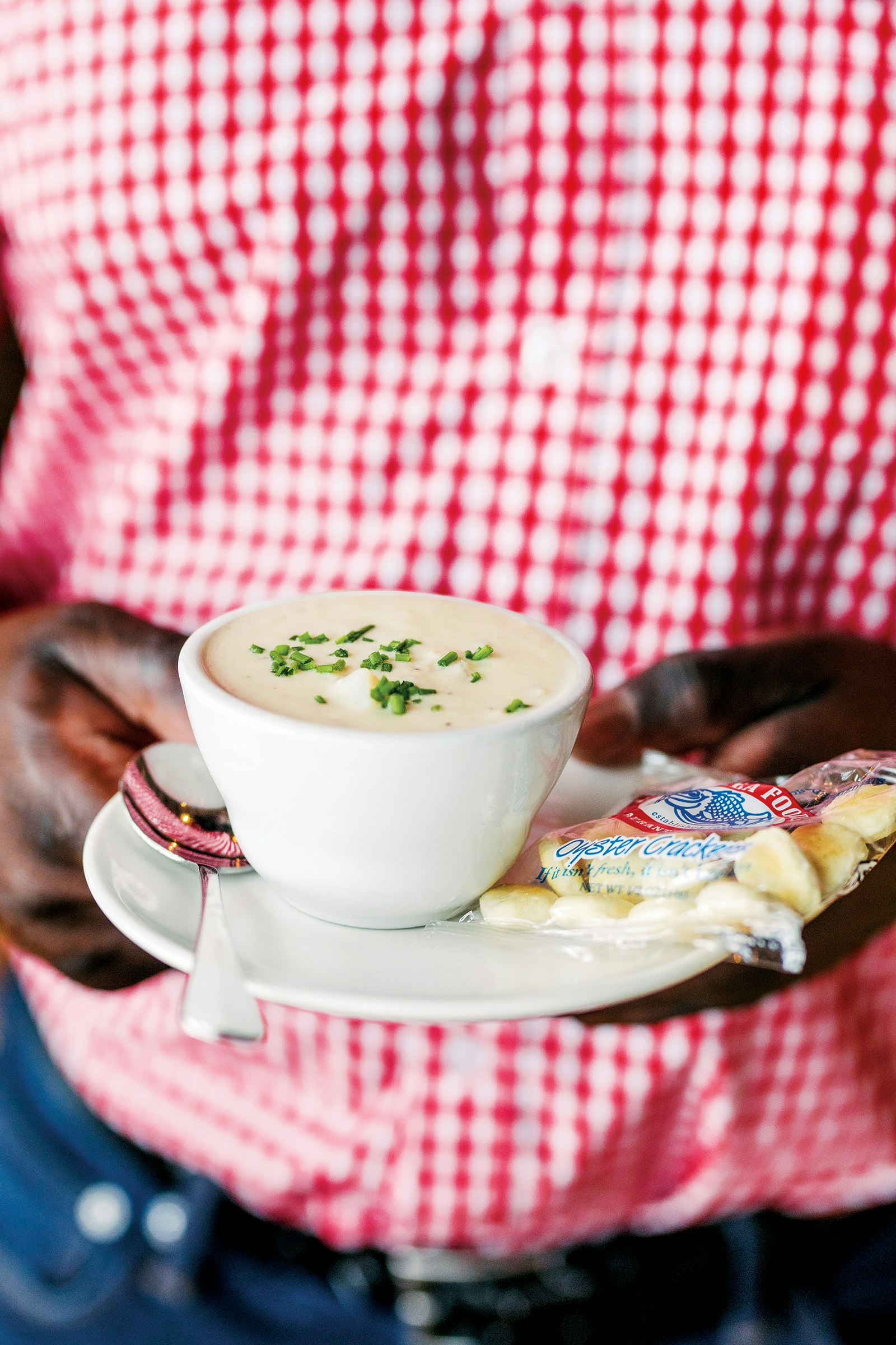 close-up shot of small white ceramic bowl of clam chowder, proffered by a man in a red gingham shirt © Adam DeTour / Lonely Planet