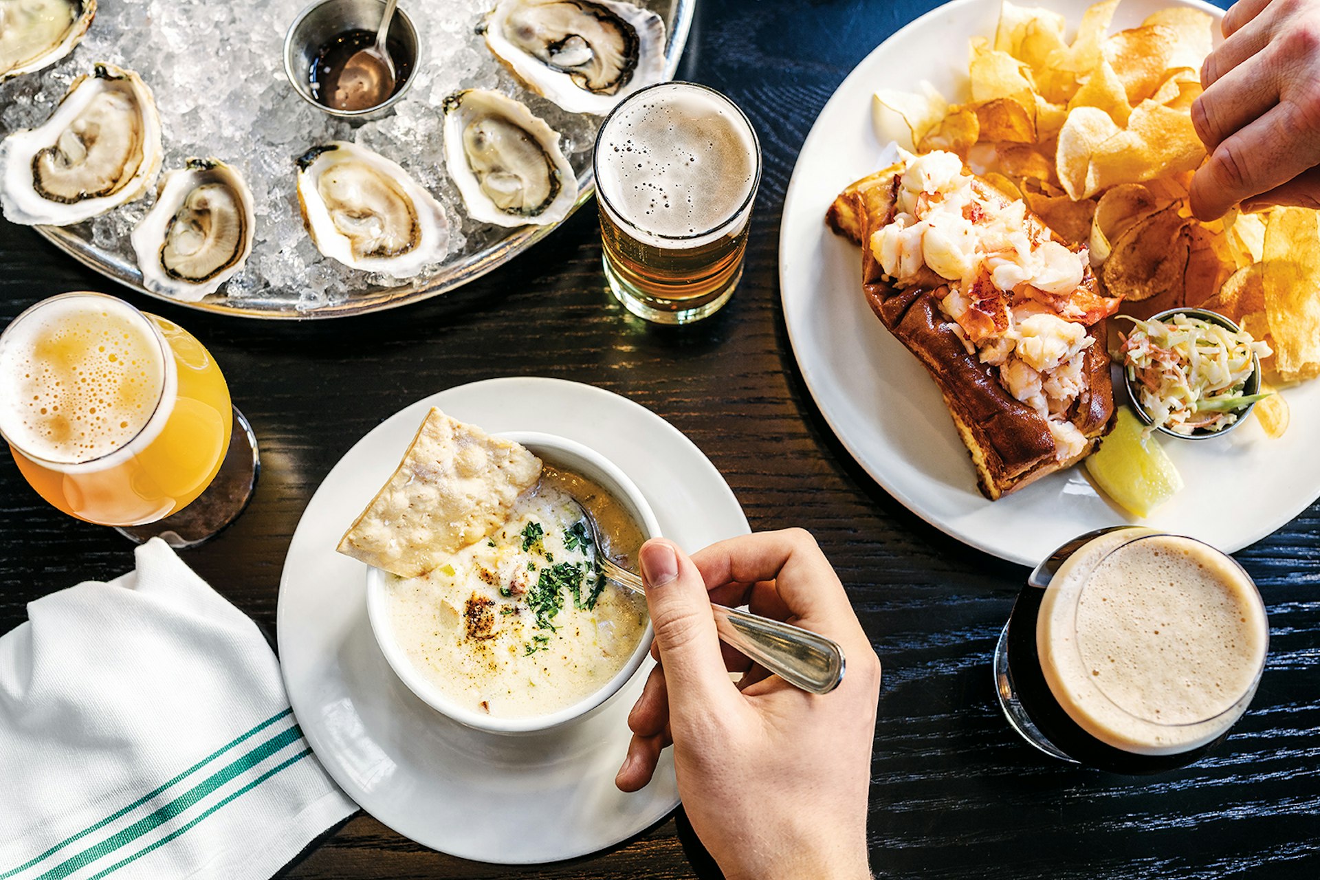overhead shot of plates of oysters, a small cup of clam chowder, a lobster roll and a beer on a dark wooden table at Row 34 in Boston © Adam DeTour / Lonely Planet