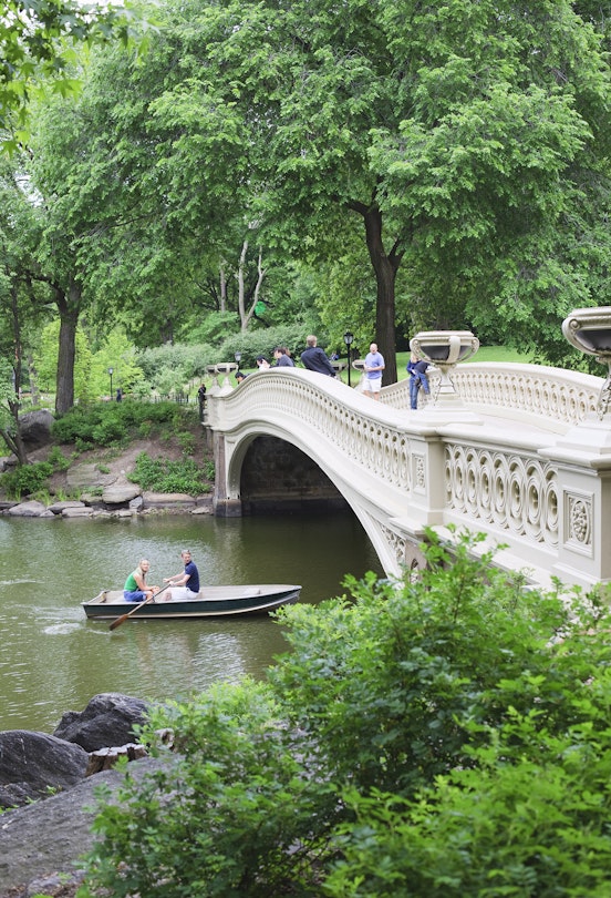 Two people row a small boat beneath Bow Bridge in Central Park in summer, as others cross the bridge on foot. © Amanda Hall / robertharding / Getty Images