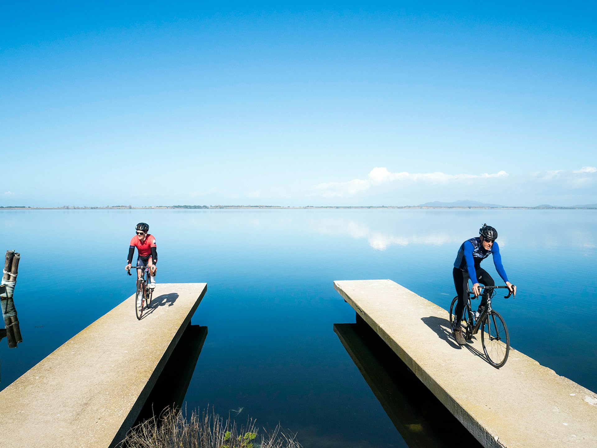 Two stark cement piers (each hosting a cyclist) reach out into the Orbetello Lagoon, which is reflecting the blue sky above © Paolo Ciaberta