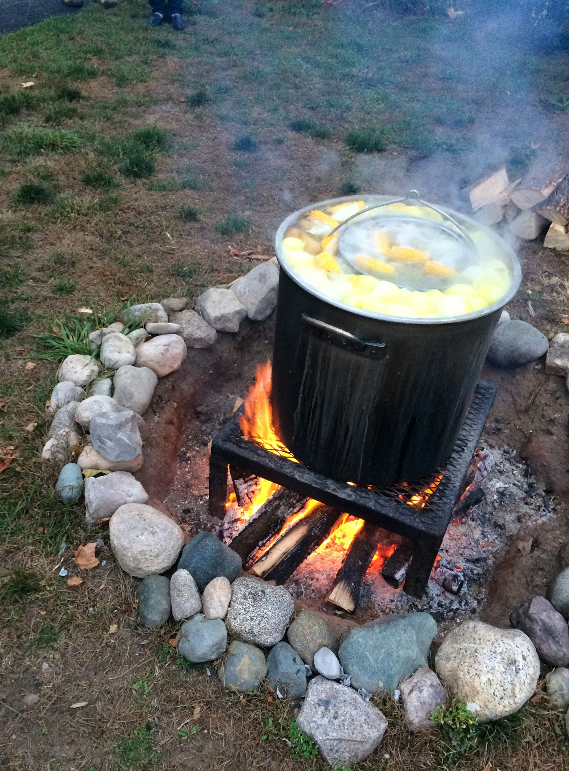 Corn floats at the top of a large cast-iron pot over an open fire outdoors as part of a fish boil