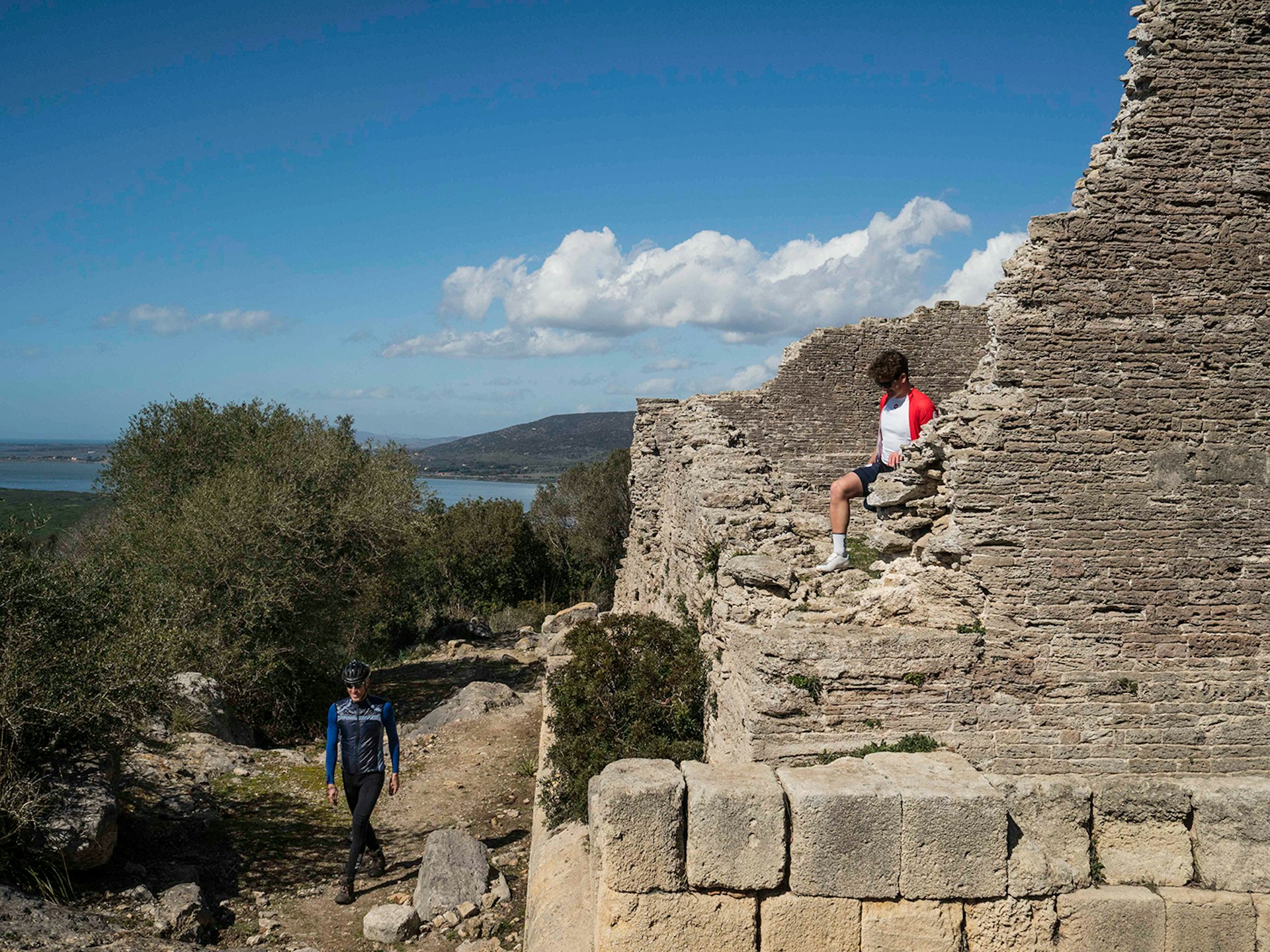 Two cyclists on foot explore the ancient Roman ruins, with the lagoon of Orbetello in the distance © Paolo Ciaberta