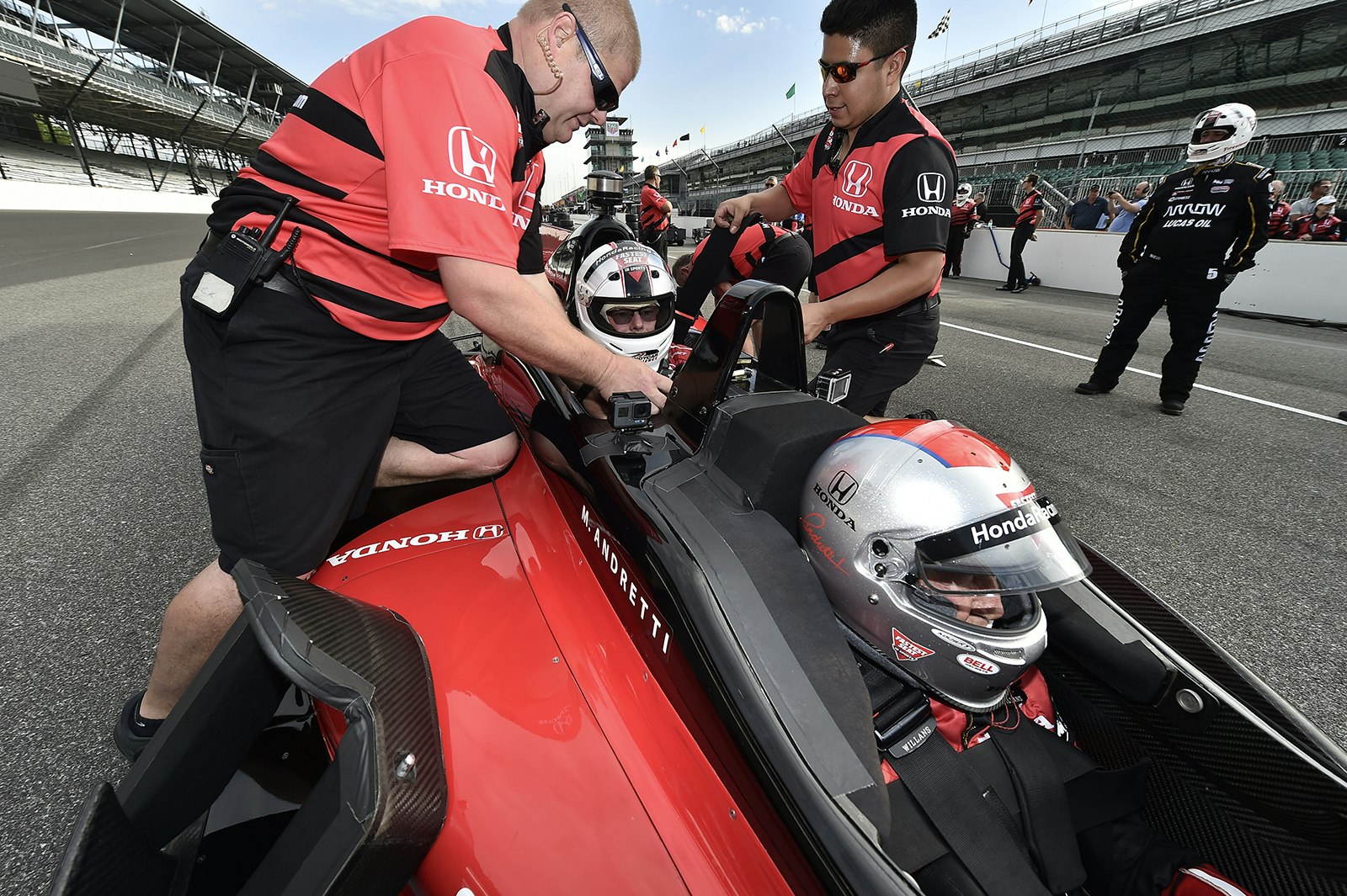 two men in red racing crew uniforms buckle in a passenger behind Mario Andretti in an IndyCar © IndyCar Digital Media