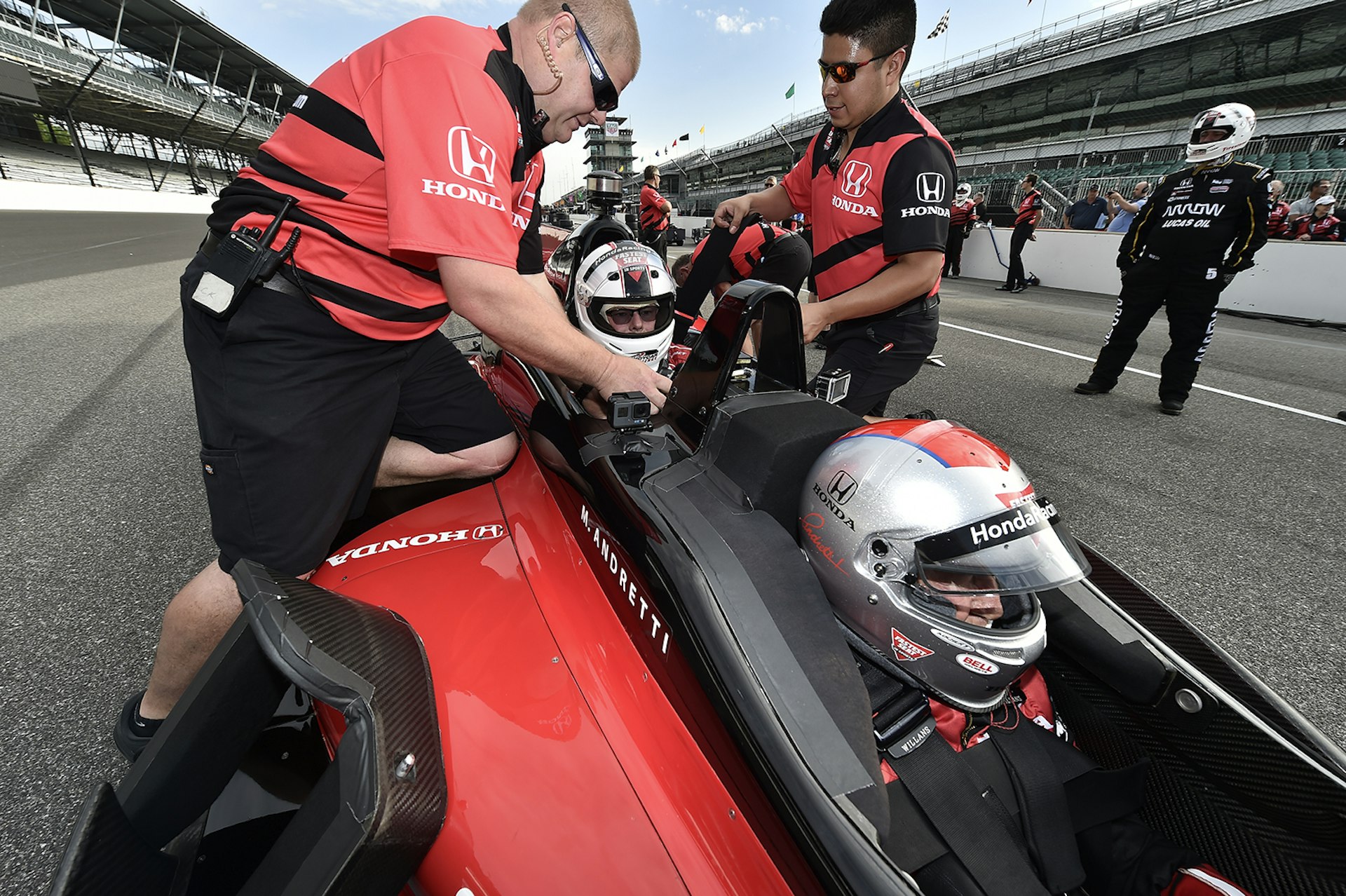 two men in red racing crew uniforms buckle in a passenger behind Mario Andretti in an IndyCar © IndyCar Digital Media