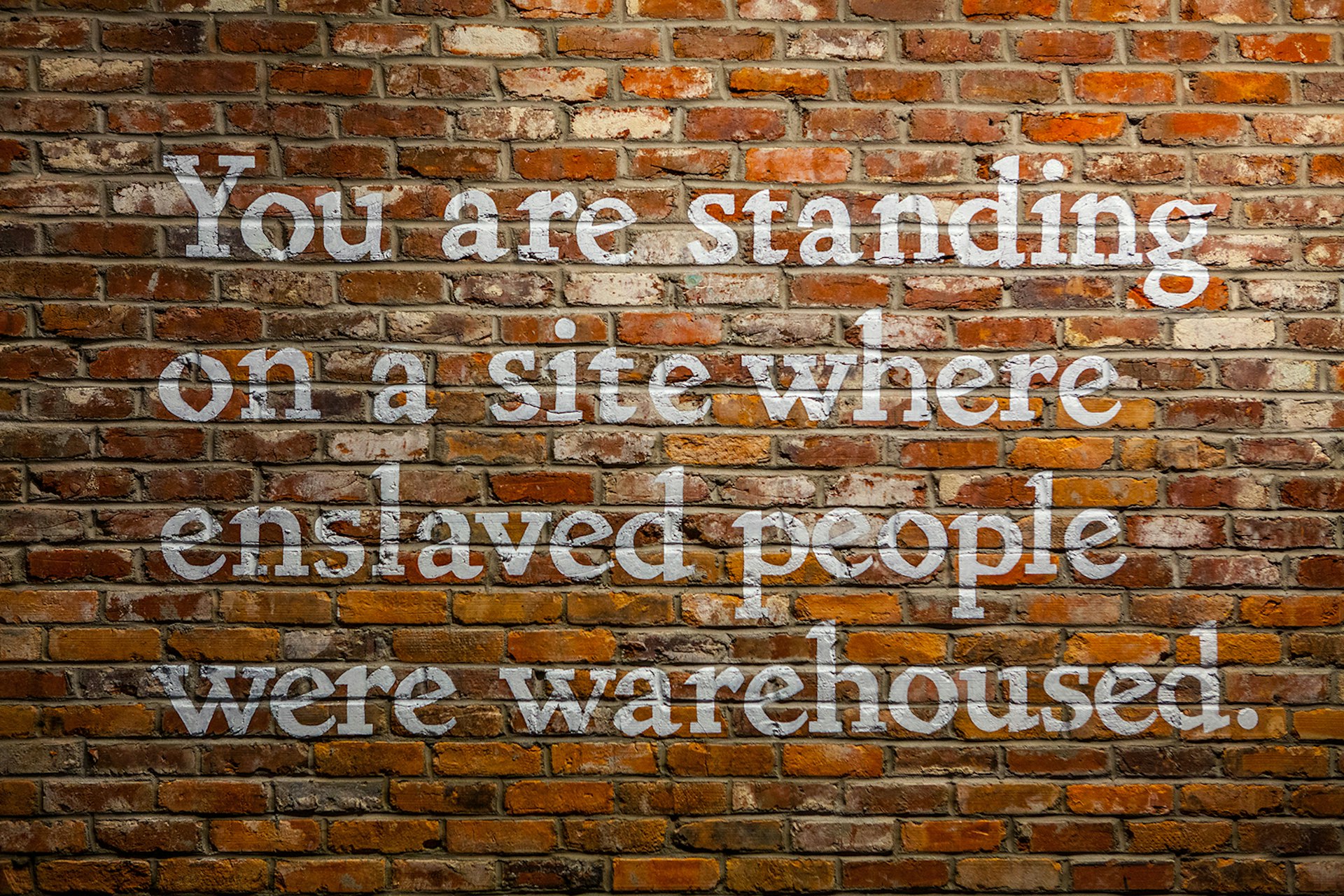 The words 'You are standing on a site where enslaved people were warehoused' is painted in white on a brick wall © Equal Justice Initiative / Human Pictures