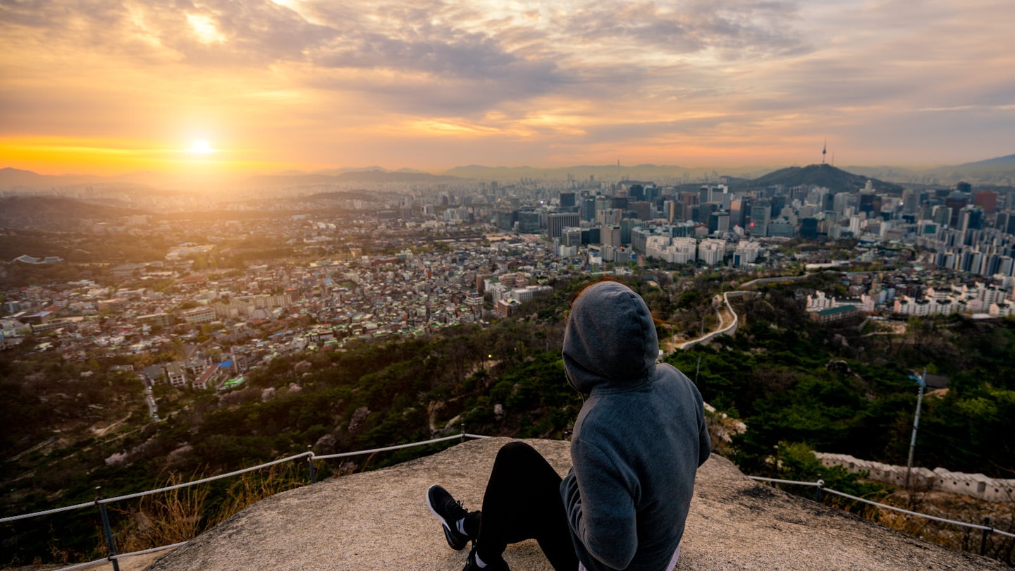 A hiker in a hoodie watches a pink and orange sunrise sitting on a rock overlooking Seoul city. Freeview: vistas of Seoul can be had via a free and gentle hike up Inwangsan © Mongkol_Chuewong / Shutterstock