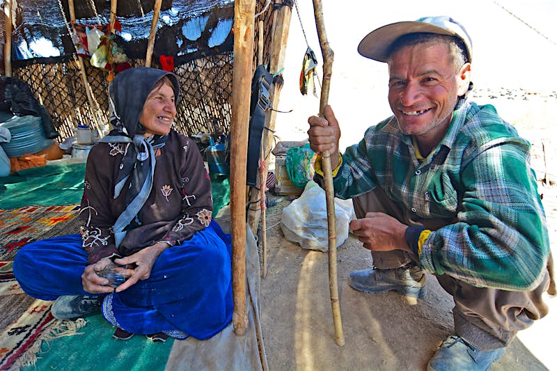Nomadic shepherds from Iran Nomadic shepherds at the Qashkai camp in the mountains of Zagros. A woman in pattered dress sits under a shelter while a man, beaming at the camera, squats next to her leaning on a walking stick.