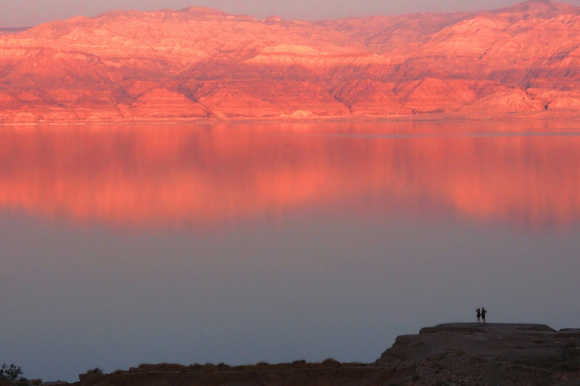 The Dead Sea glowing orange in the sunset, with the outline of two figures visible on the nearest bank © GMa / Shutterstock