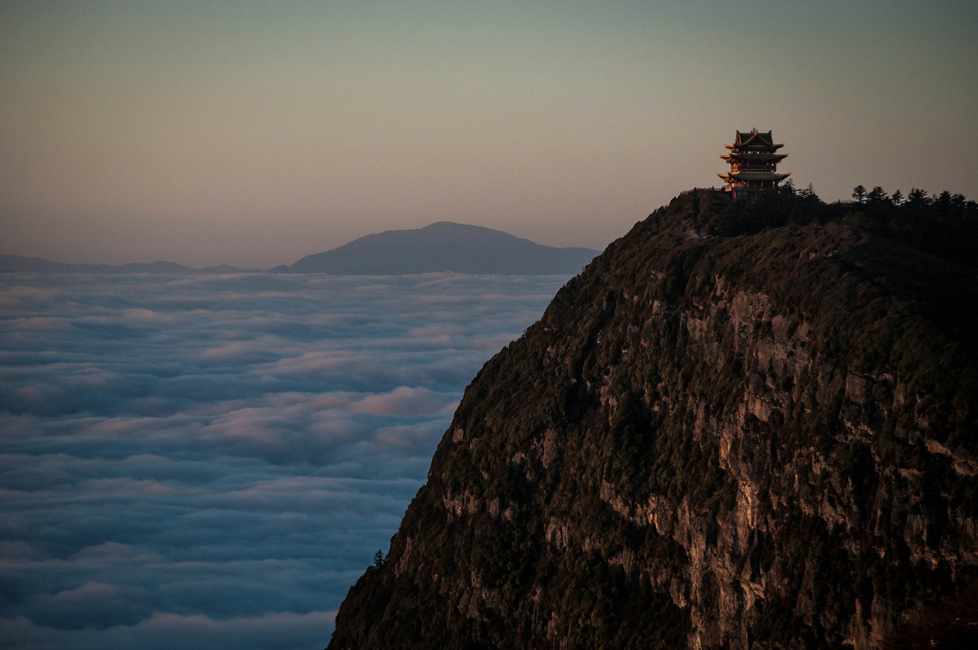 The summit of Éméi Shān above the cloud line. A lone temple stands near the summit, cast into shadow by the setting sun.