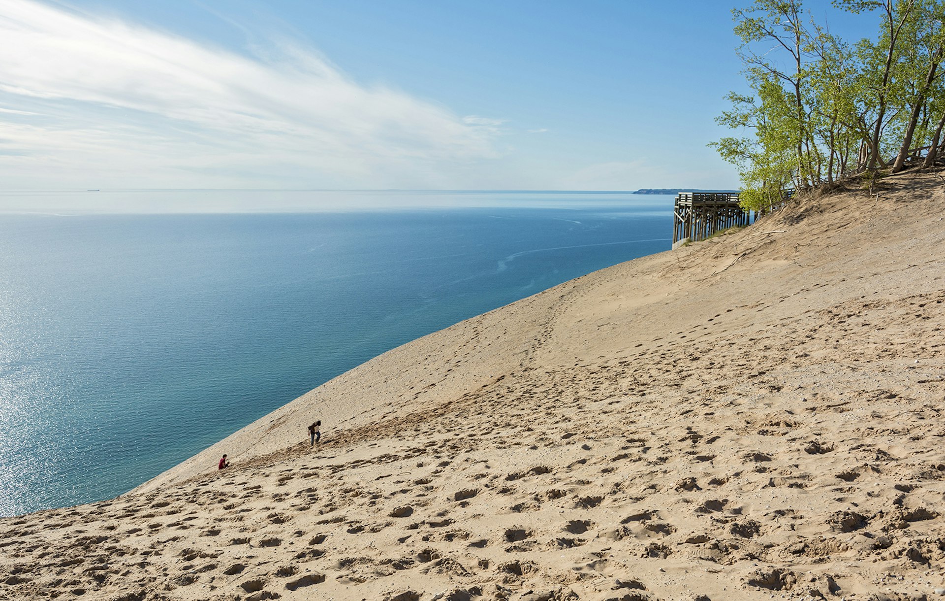 Two people hike up a massive golden sand dune with Lake Michigan in the background
