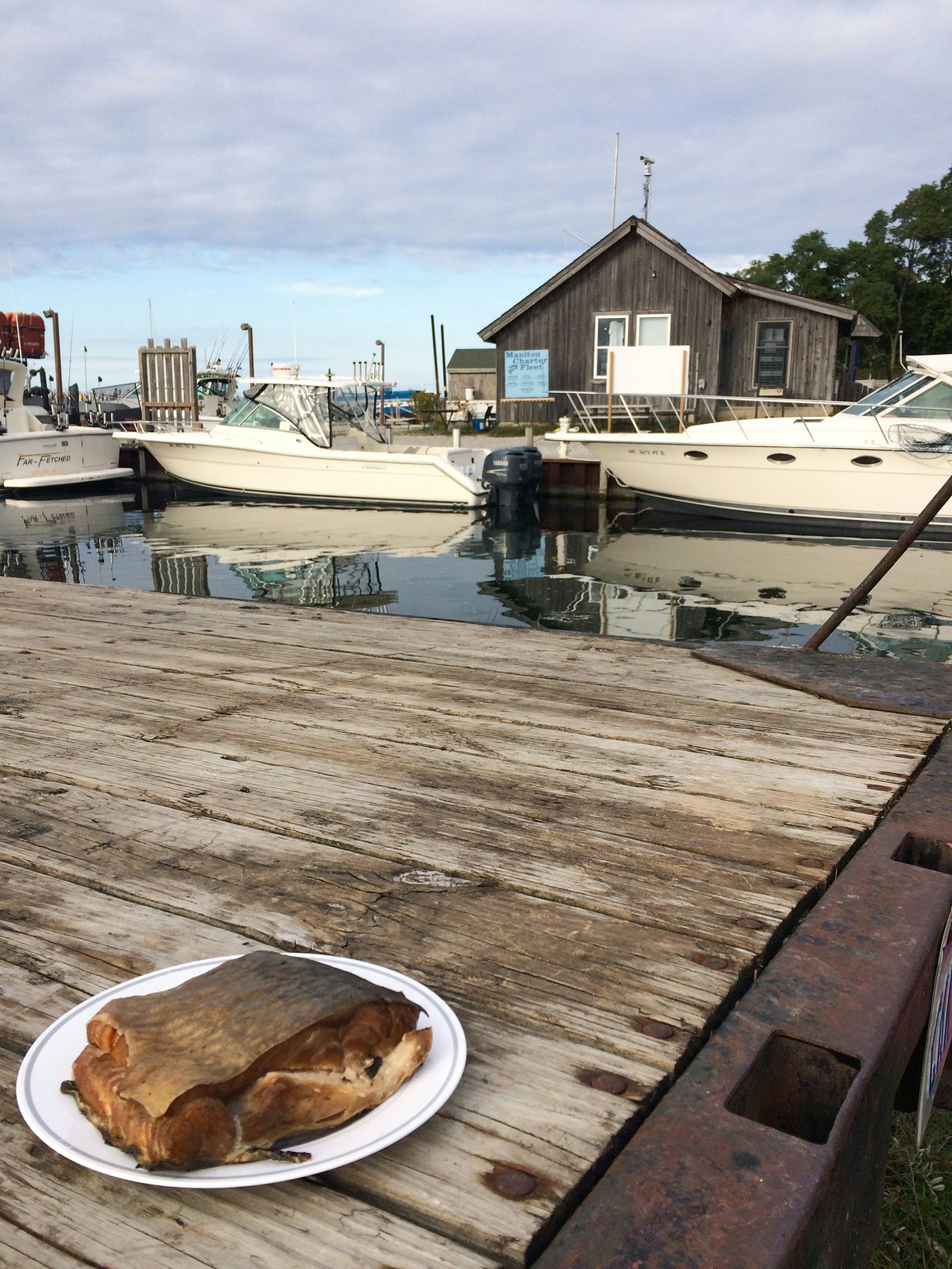 A plate of smoked fish sits on a dock with boats and fishing shanties in the background