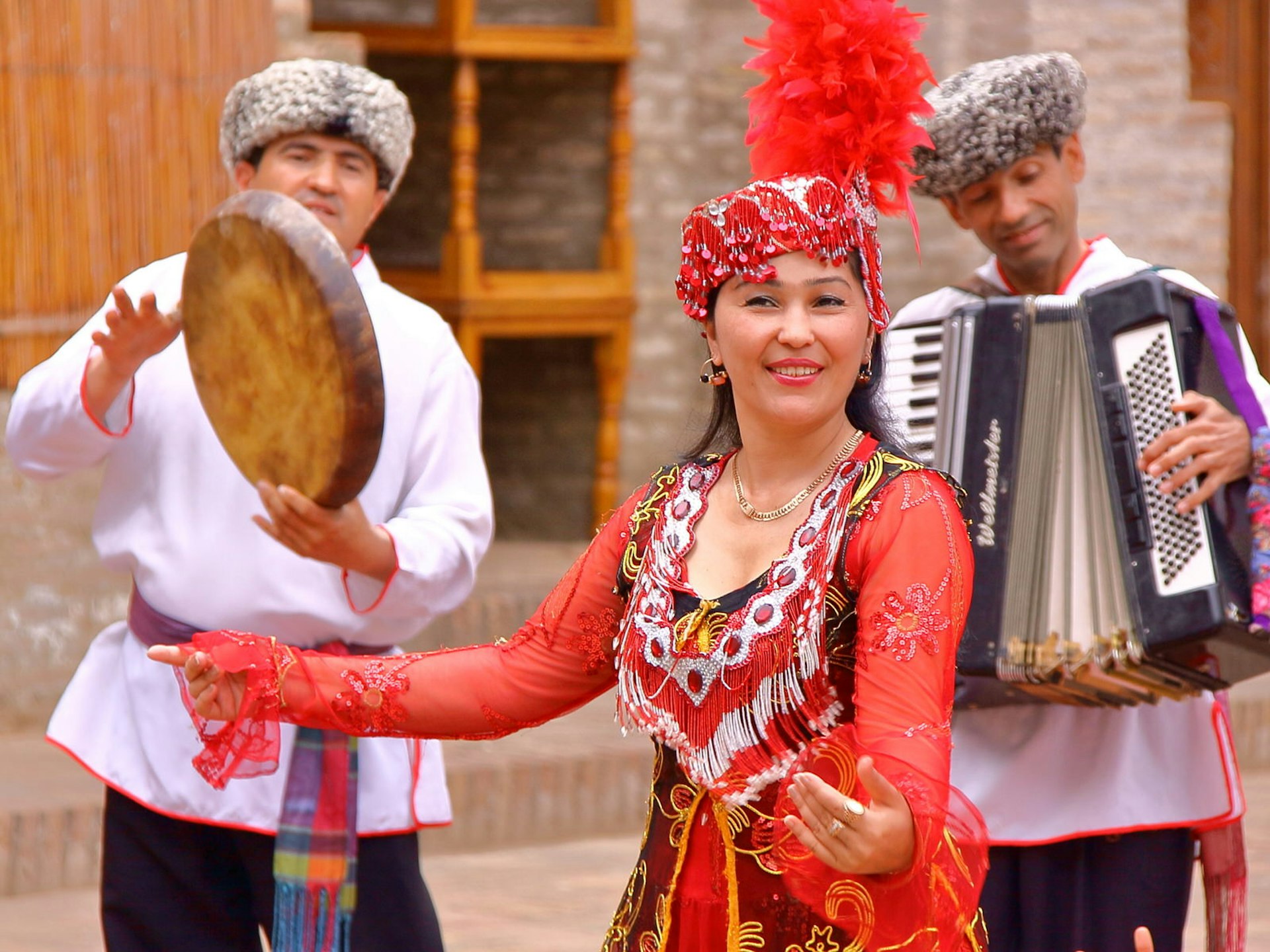 A woman in red traditional Uzbek costume with beads and a feathered hat dances in front of two musicians, one drummer and one accordion player. Welcoming people: performers demonstrate traditional Uzbek music and dance in the ancient city of Khiva, soon to be connected by high-speed rail © Christophe Cappelli / Shutterstock