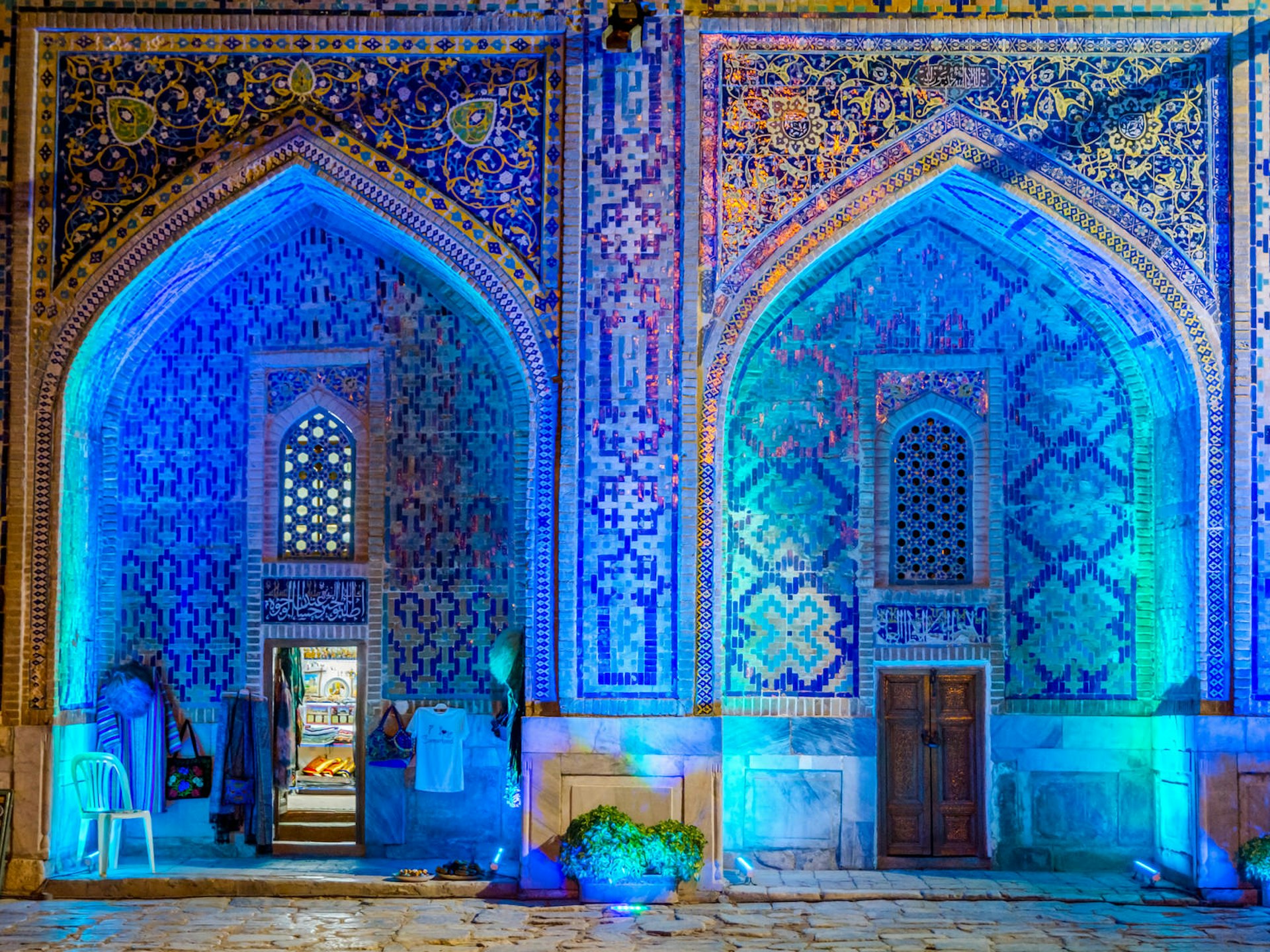 Two blue tiled arches are illuminated with glowing blue and green lights. Shop in lit up at night in the atrium of Sher Dor medressa in Samarkand © Dinozzzaver / Shutterstock