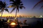 A long-distance view of the Miami skyline is seen over a dark bay at sunset. In the foreground is an outdoor bar area with palm trees and a fire pit © Louis Davilla / Getty Images