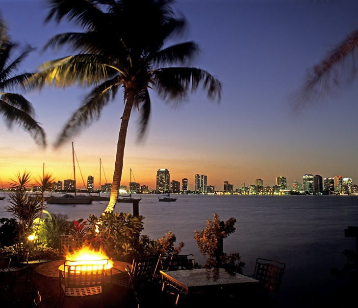 A long-distance view of the Miami skyline is seen over a dark bay at sunset. In the foreground is an outdoor bar area with palm trees and a fire pit © Louis Davilla / Getty Images