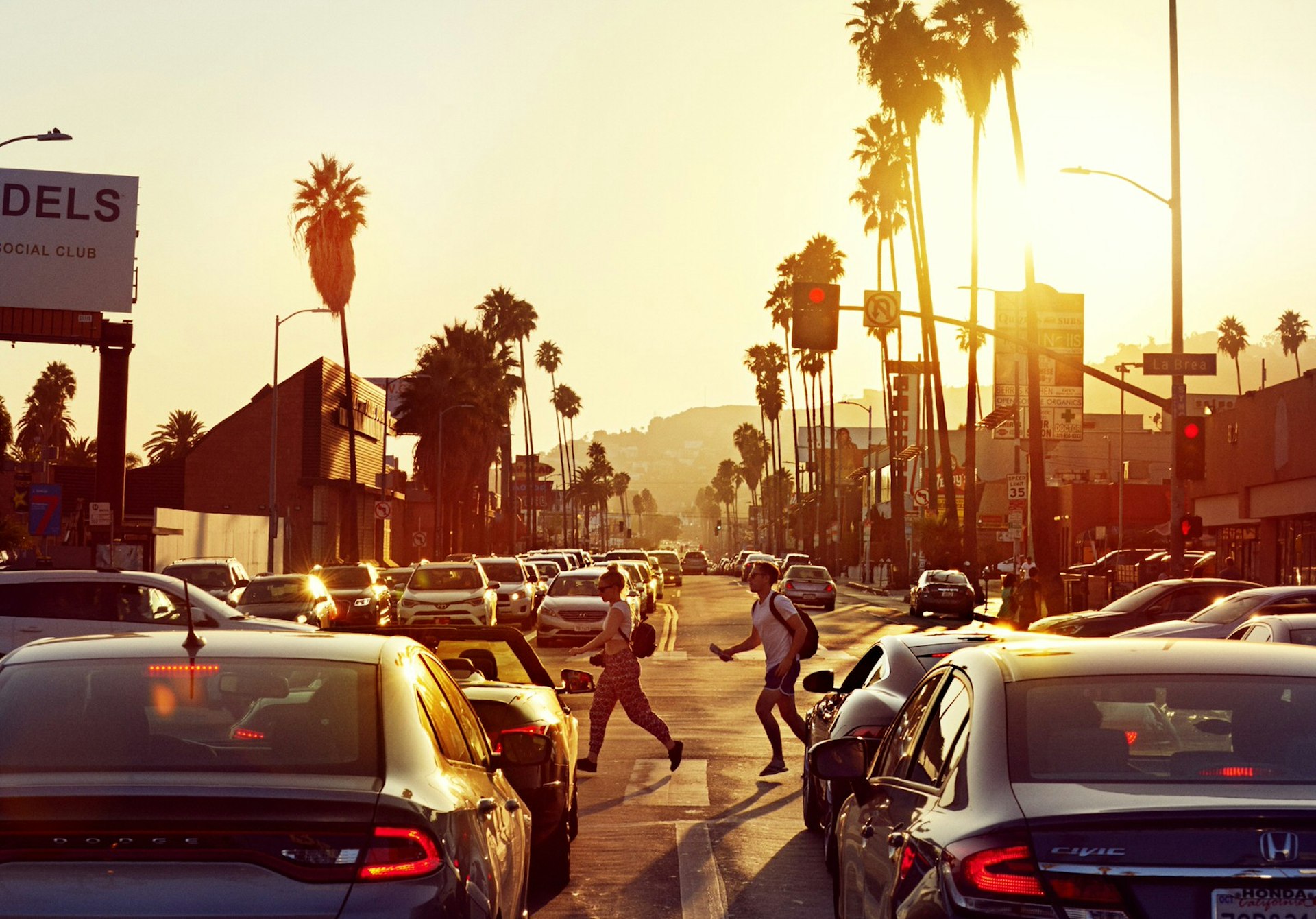 A couple of pedestrians cross a busy LA road at sunset.